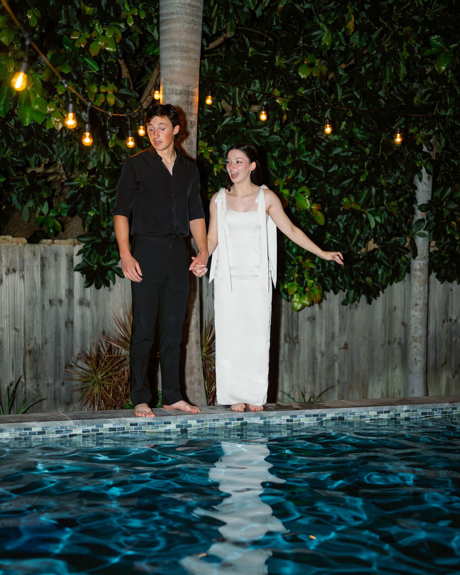 Couple standing near illuminated pool at night with string lights overhead.