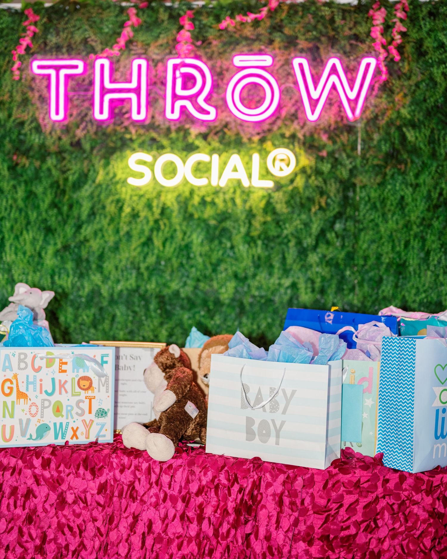 Colorful neon signs illuminate a display of wrapped gifts and pink decor against a green hedge backdrop.