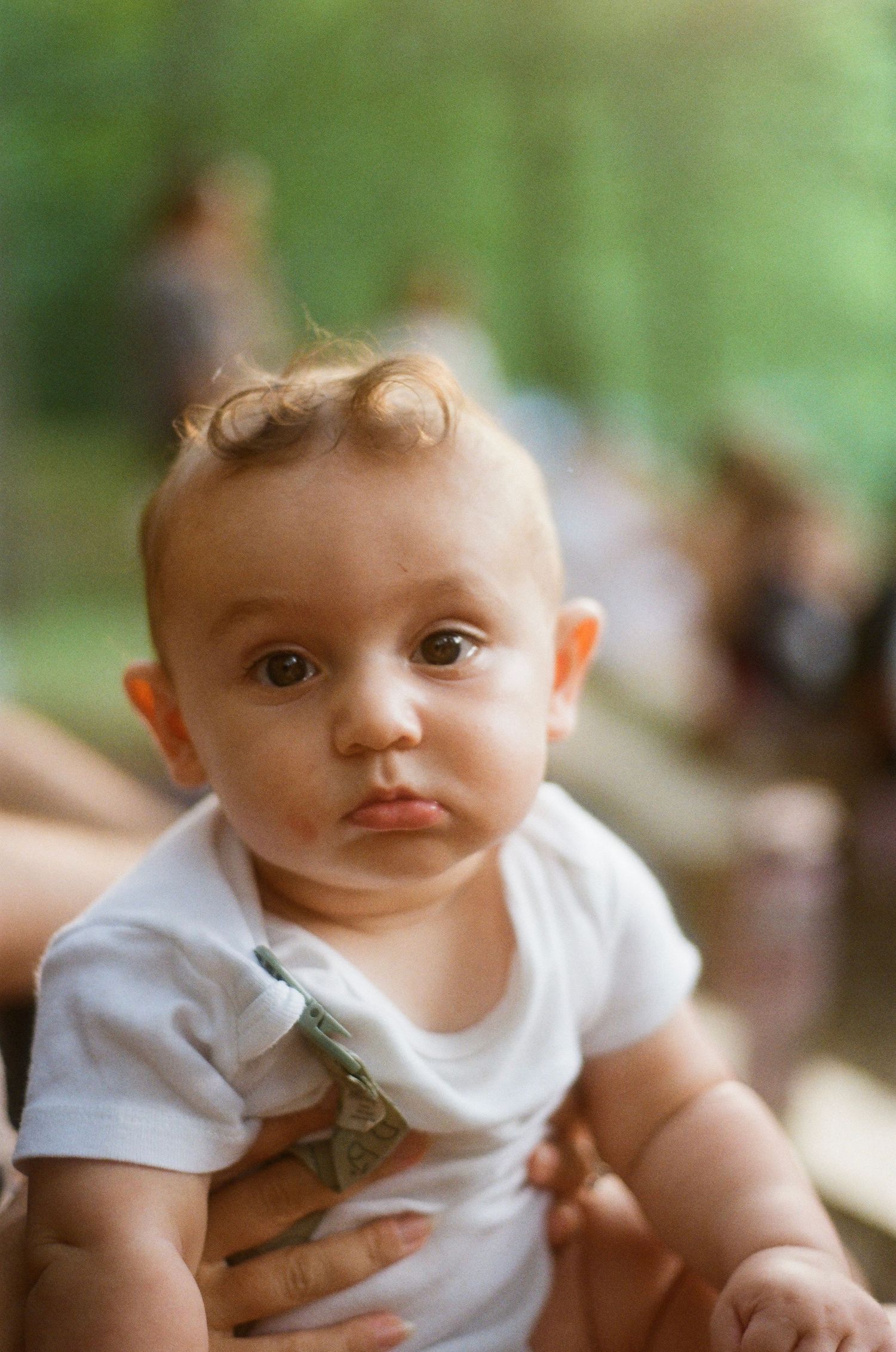 A cute baby in a white onesie with a pouty expression being held outdoors with a blurred green background.