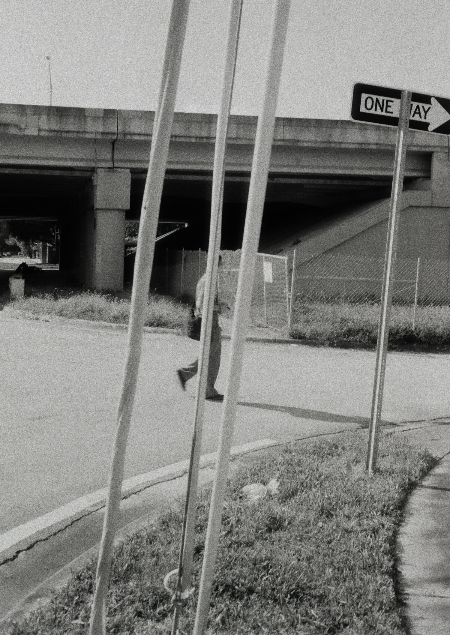 Black and white photo of a person walking near an overpass with a one-way street sign and chain-link fence.