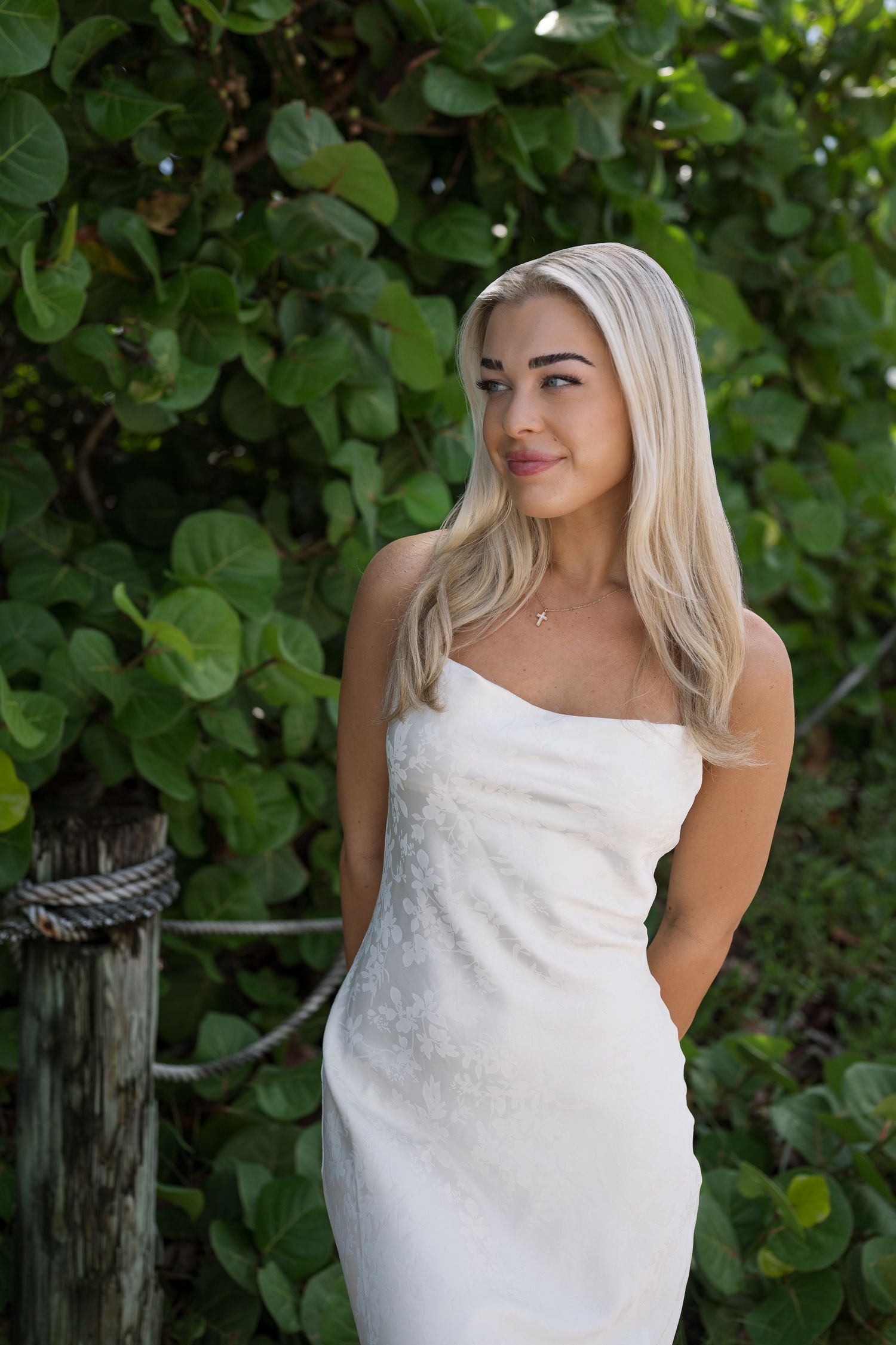 Woman in white sleeveless dress poses against green foliage background in outdoor portrait series.