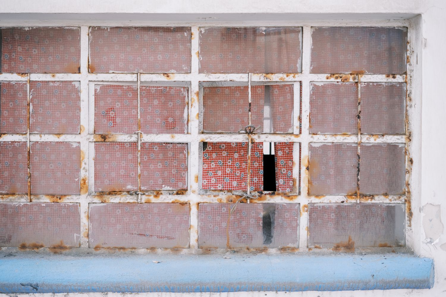 Rusty deteriorating window frame with peeling paint and floral wallpaper visible through broken glass panes.