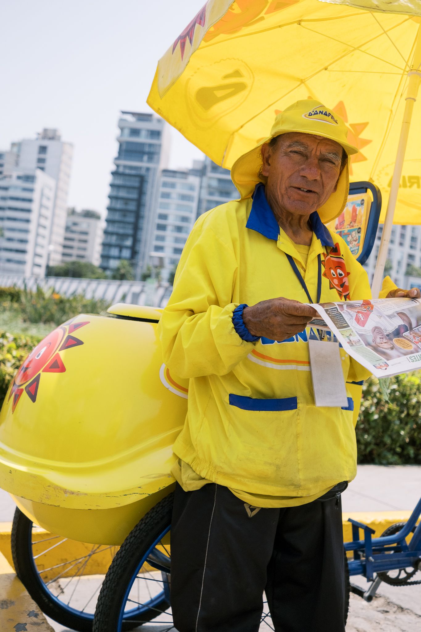 Elderly street vendor in yellow uniform and hat selling items next to a yellow cart under a yellow umbrella outdoors.