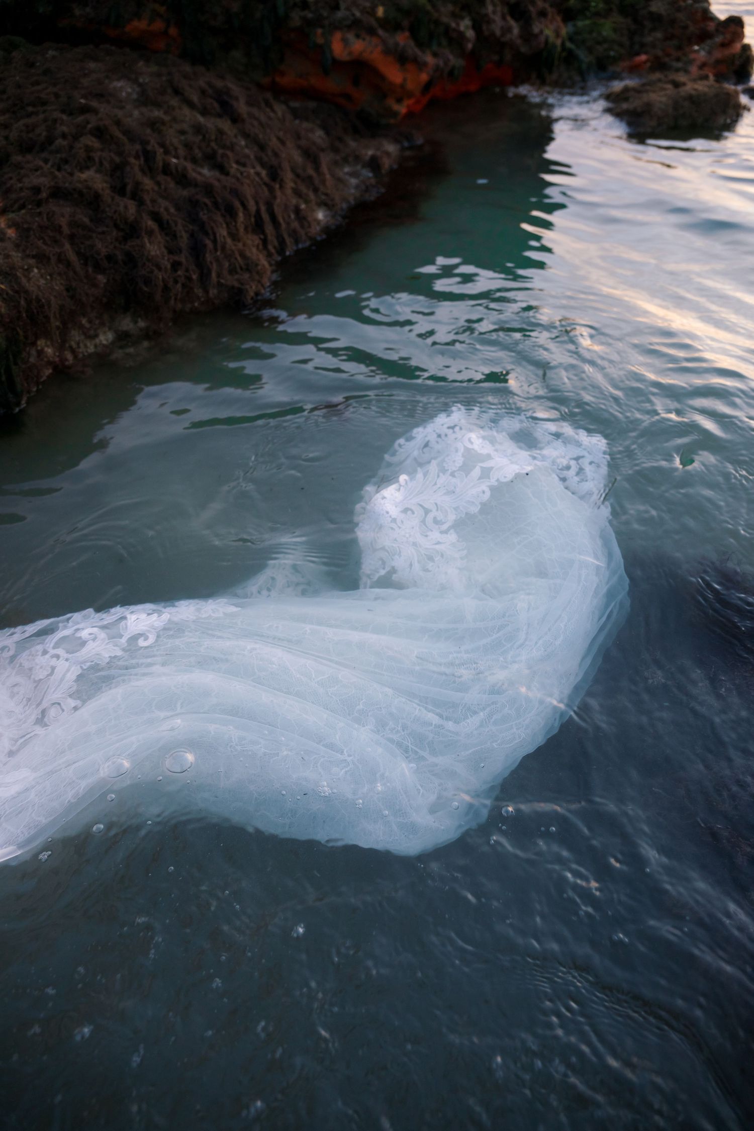 White wedding dress fabric flows dramatically in turquoise ocean waters against rocky cliffs.
