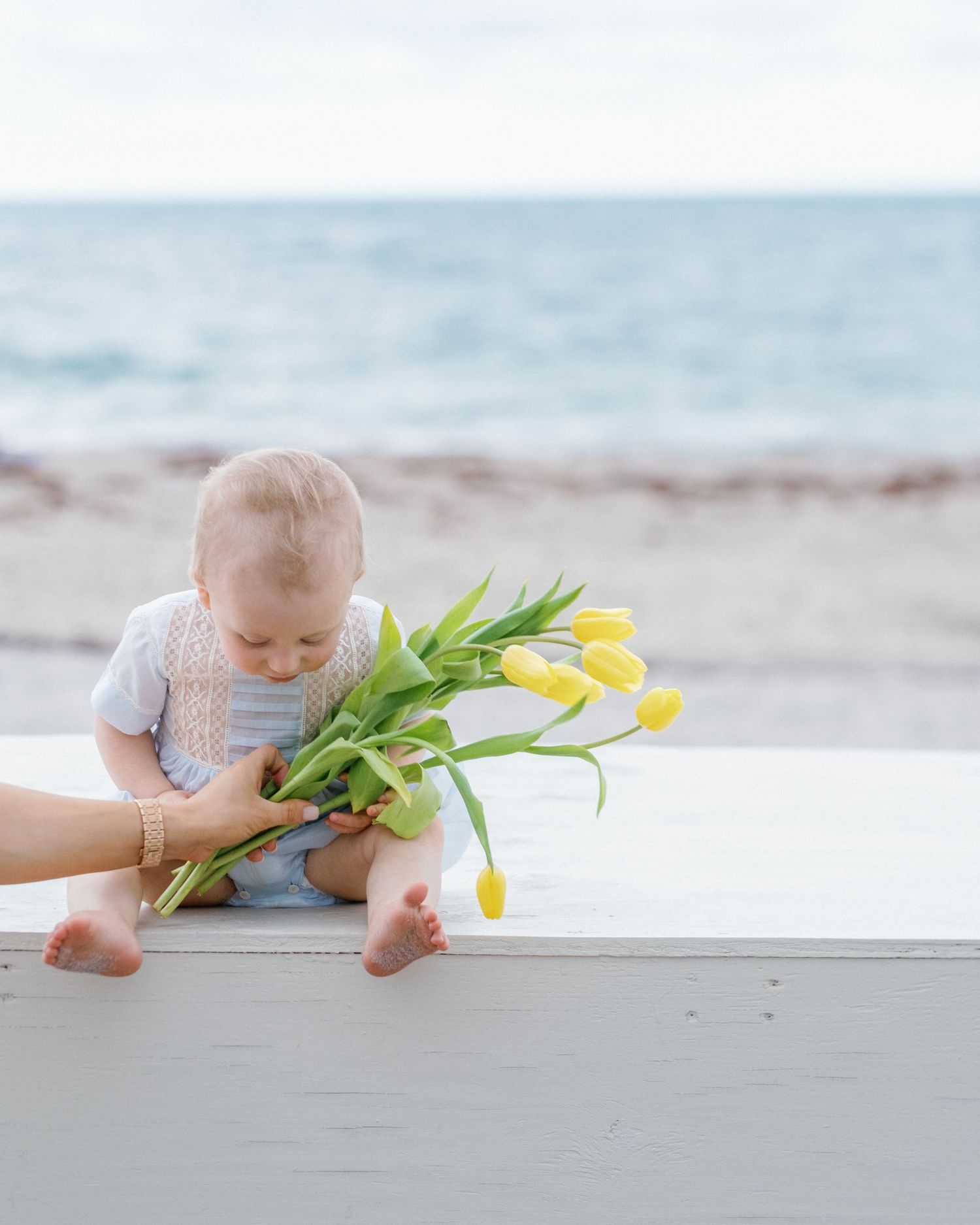 A baby lies on a white surface reaching for yellow tulips in a soft, beachy setting.