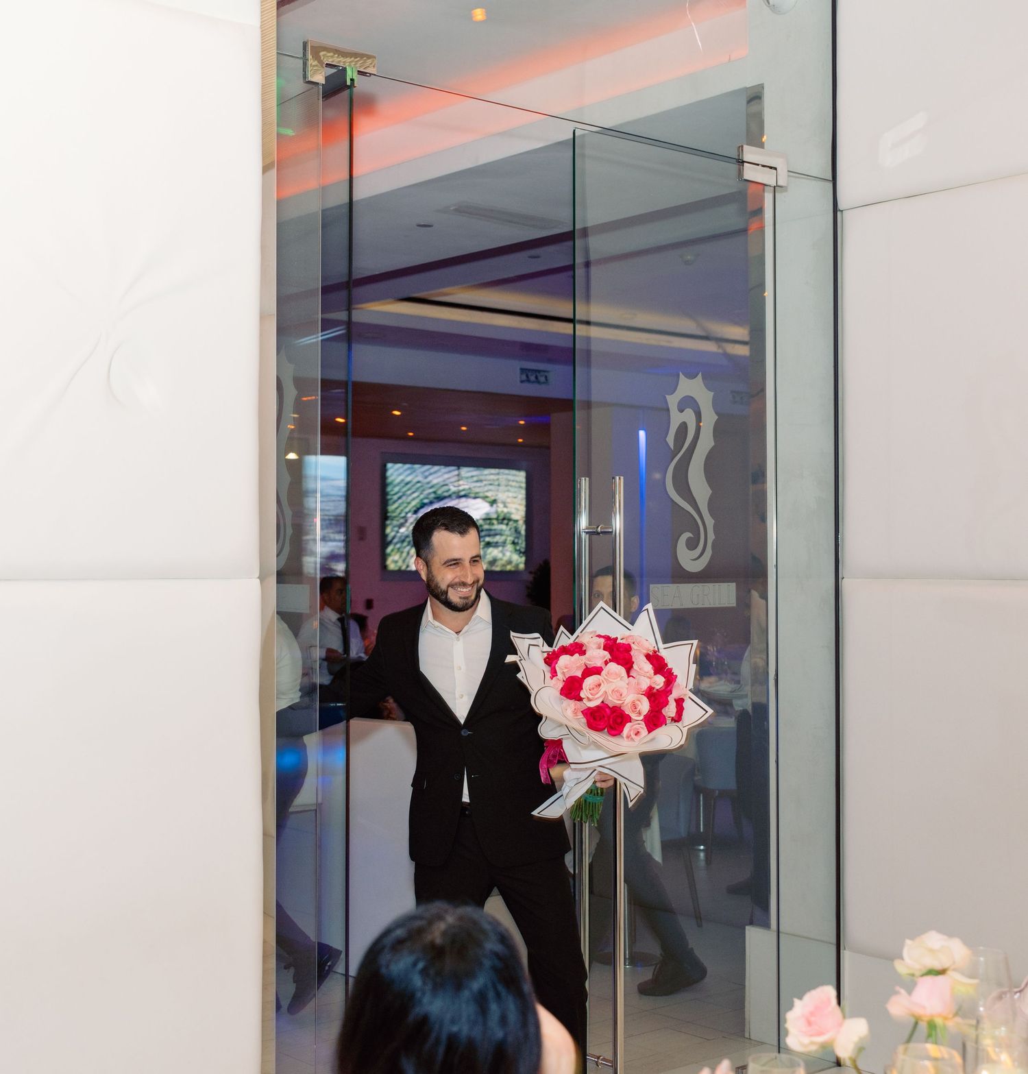 A person in a dark jacket carries pink flowers through a modern glass-walled space with white and gray decor.