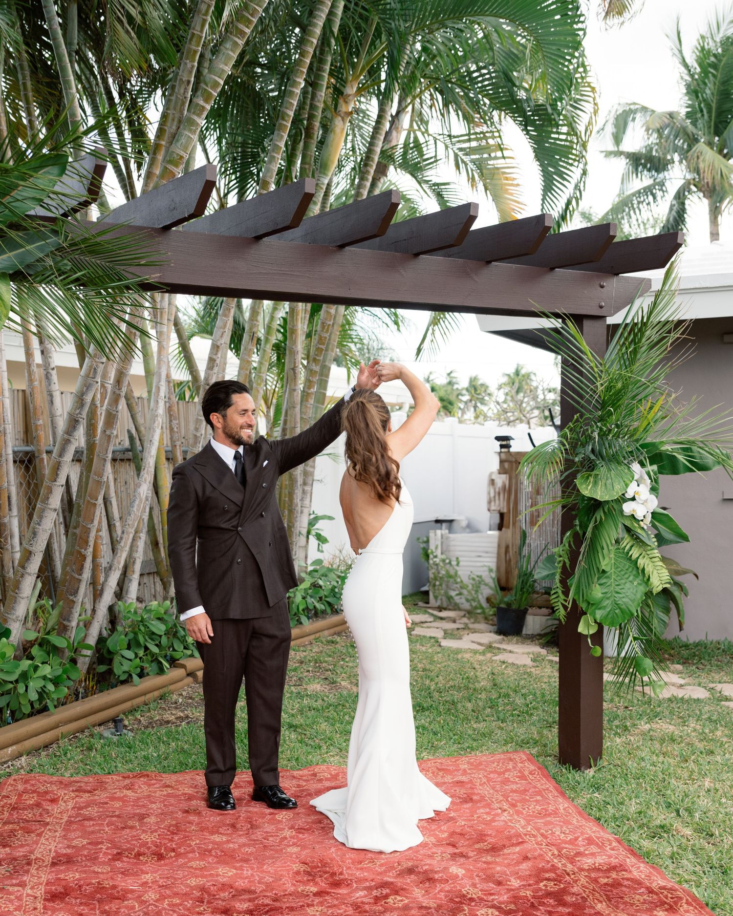 A couple shares a romantic moment under a wooden pergola with palm trees in a backyard garden setting.