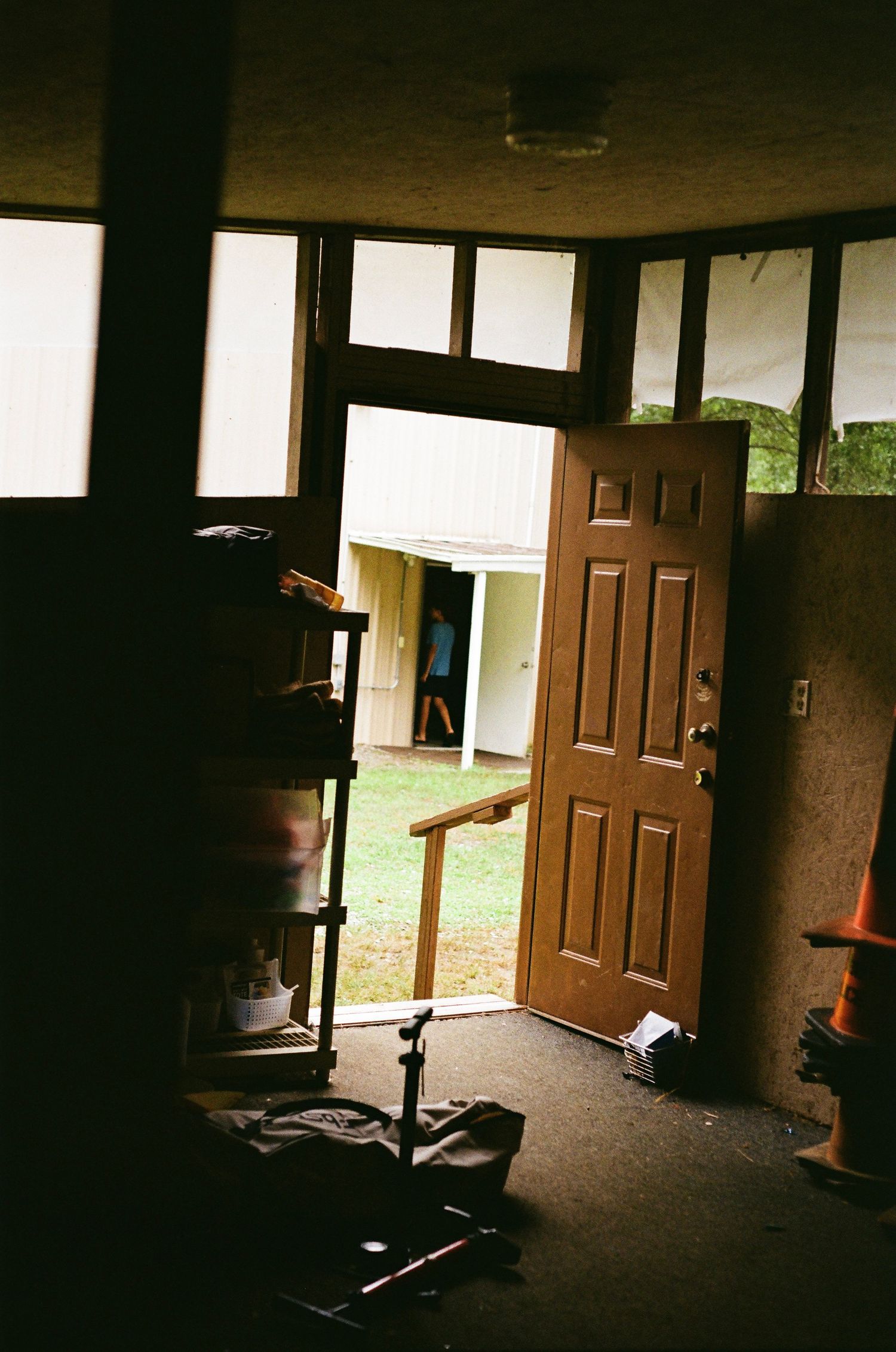 Interior view of an open wooden door leading to a green yard, with cluttered room items visible in dim lighting.