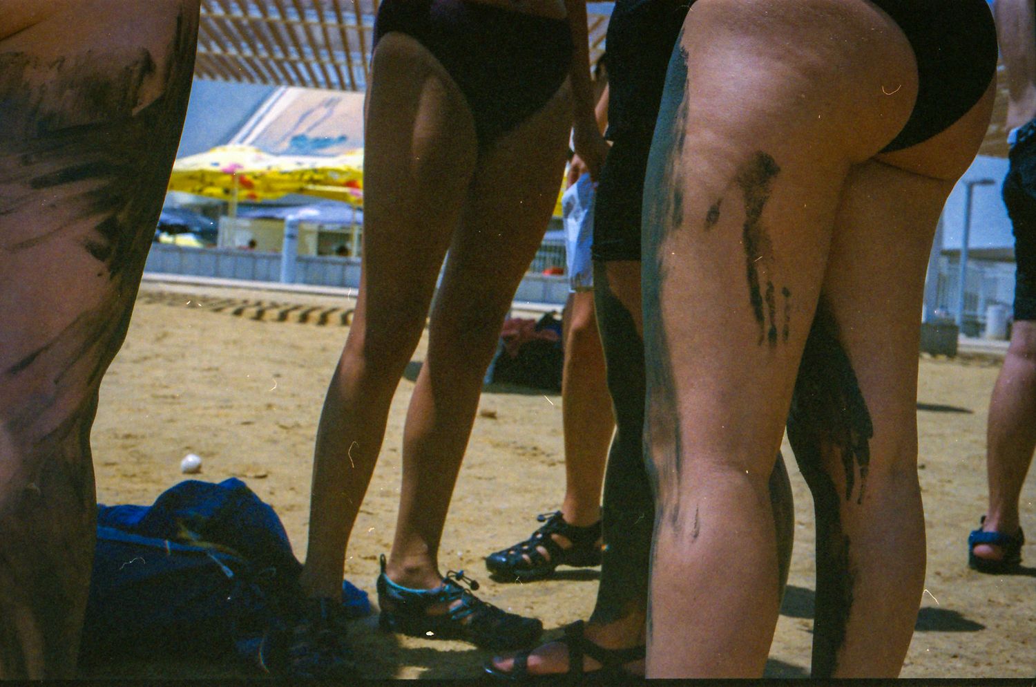 People standing on sandy beach in swimwear, legs covered in dark mud or paint, with bags on ground nearby.