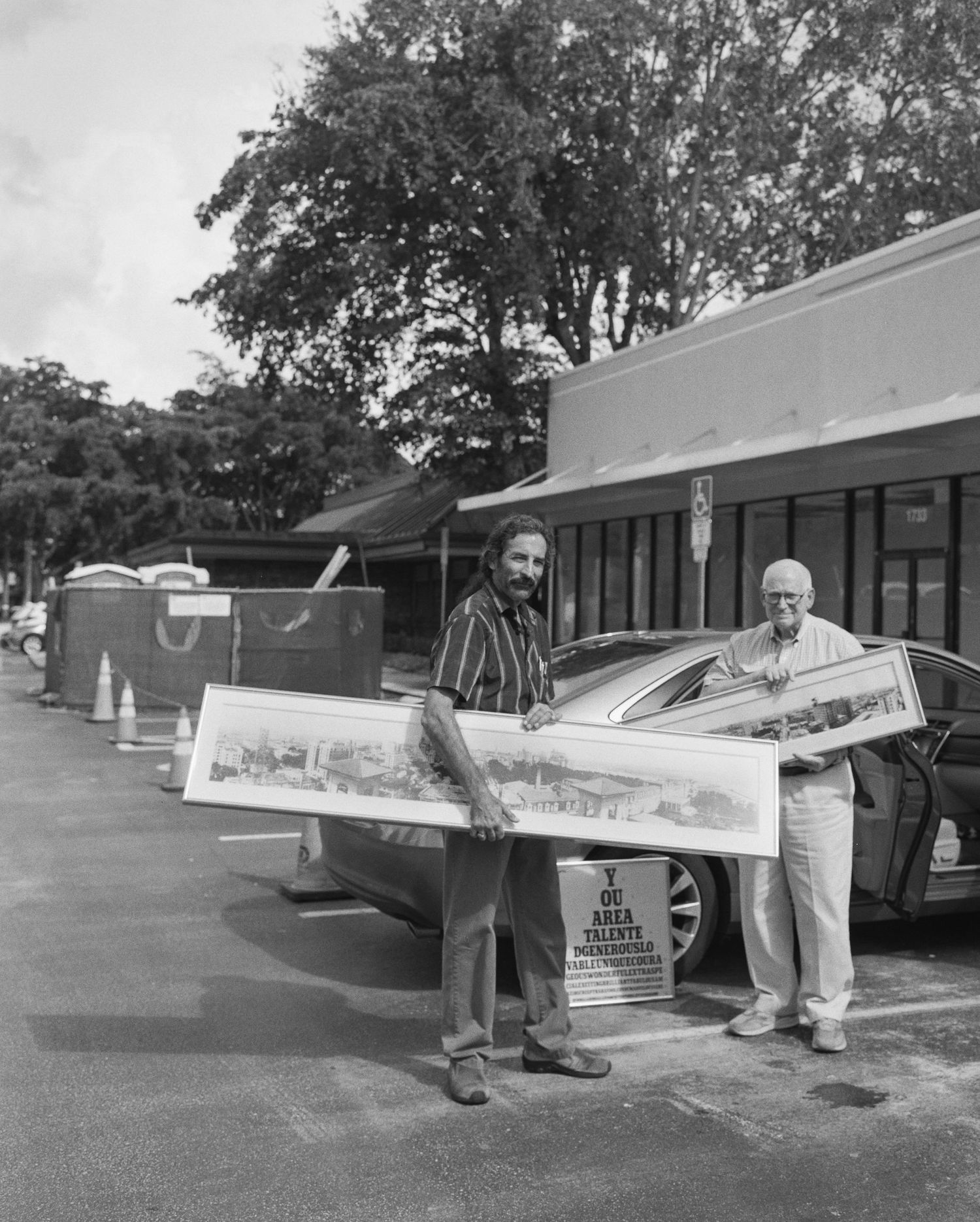 Two men holding large panoramic artwork prints outdoors near a parking lot and storefront in a black and white photo.