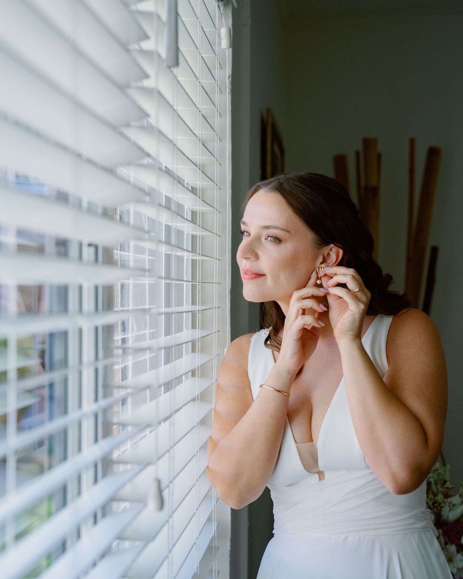 Person in white top adjusts earrings while looking through window blinds in moody lighting.