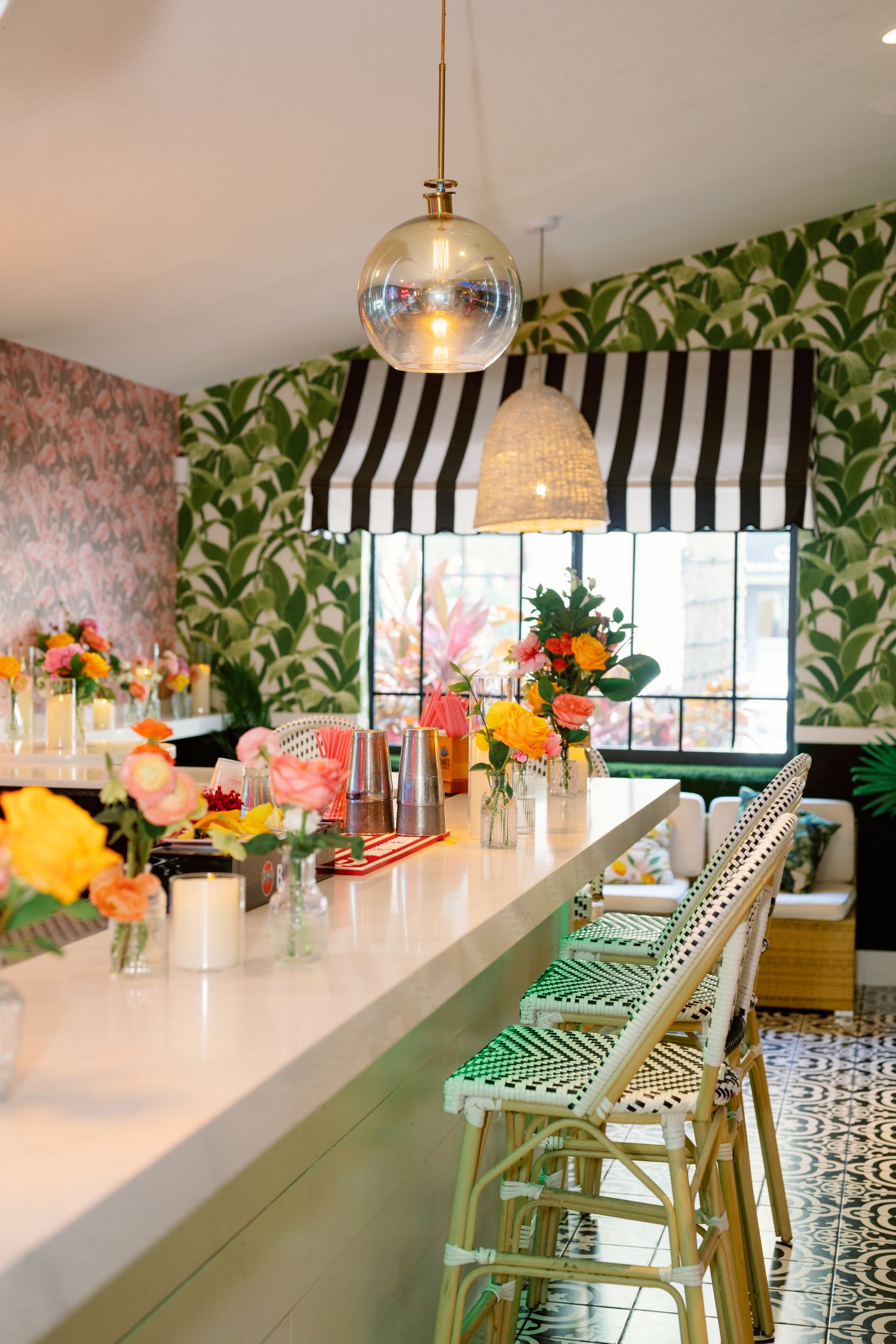 A modern tropical-themed bar with palm leaf wallpaper, striped awnings, and colorful floral arrangements.