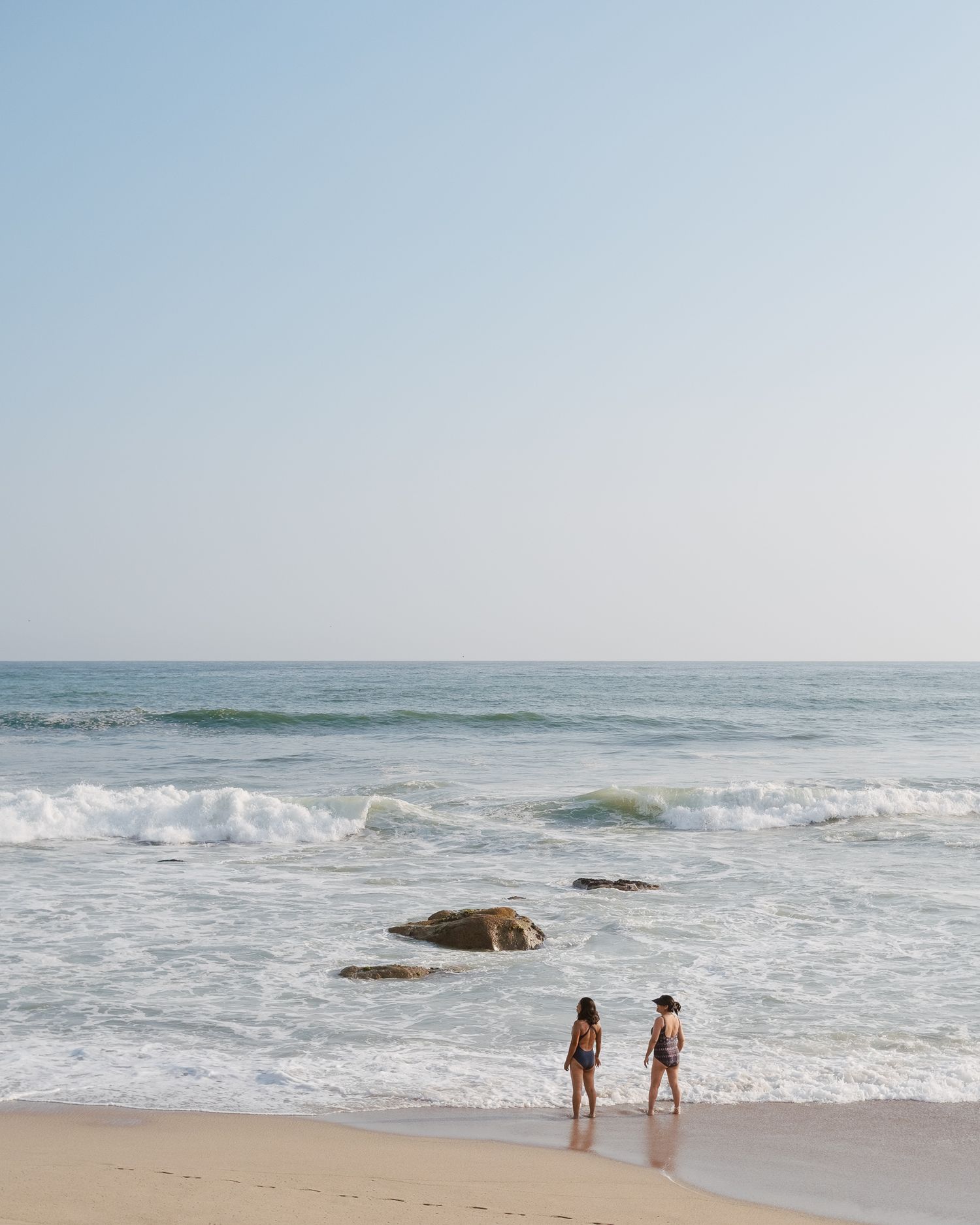 Two people walking along a scenic beach shoreline with crashing waves and rocks visible in the ocean.
