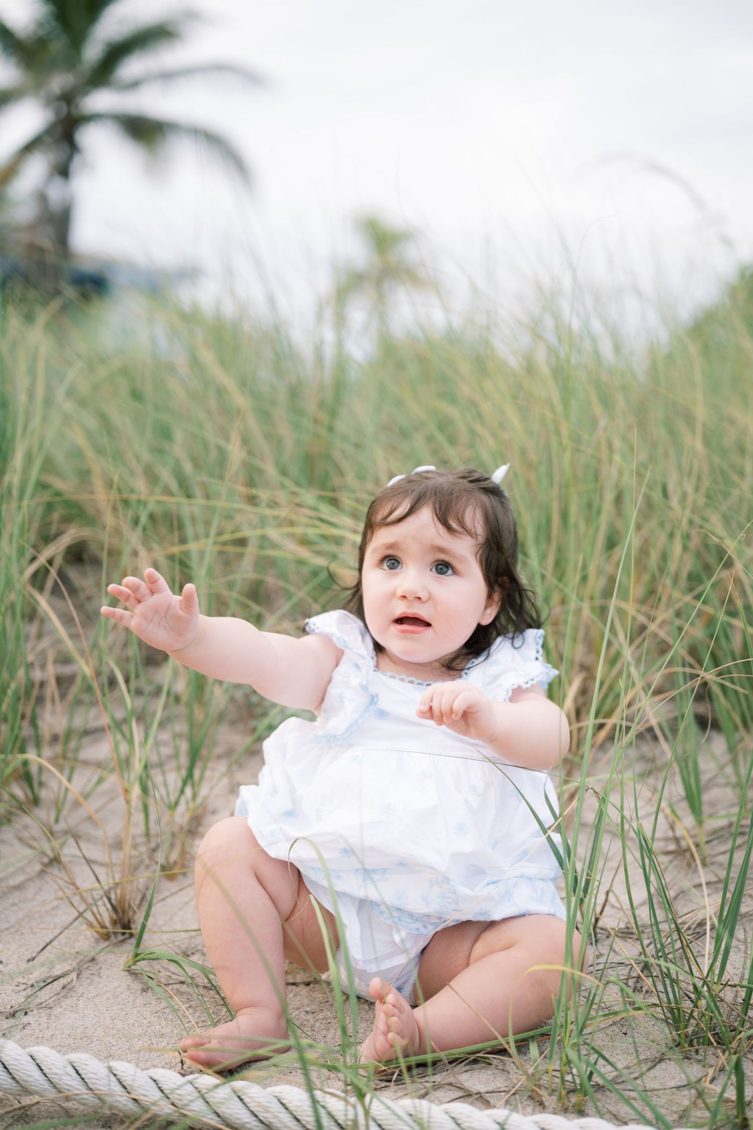 A baby in a white dress plays joyfully on a sandy beach with sea grass on a cloudy day.