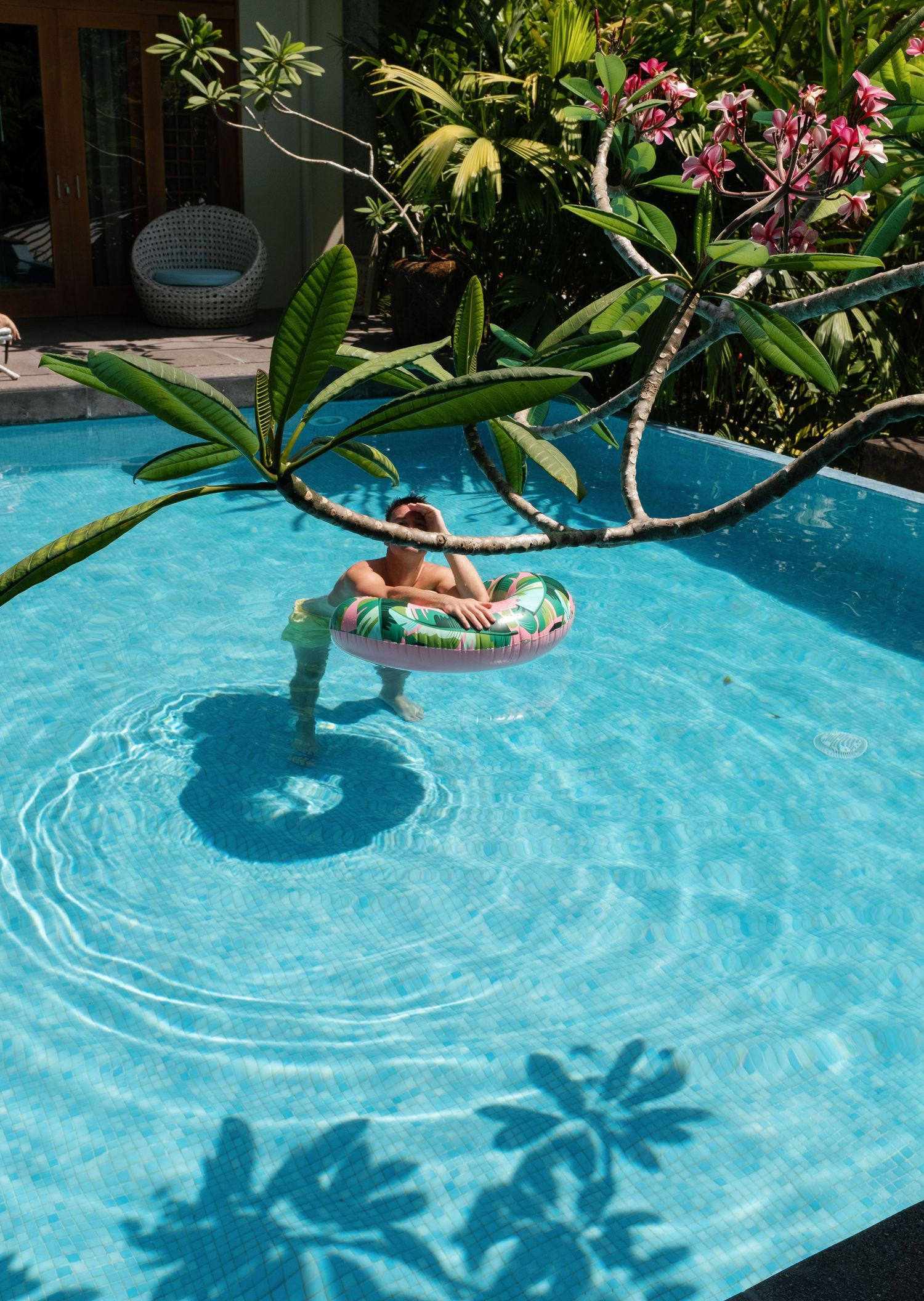 Someone floats in a bright turquoise pool with tropical plants casting shadows on the water surface.