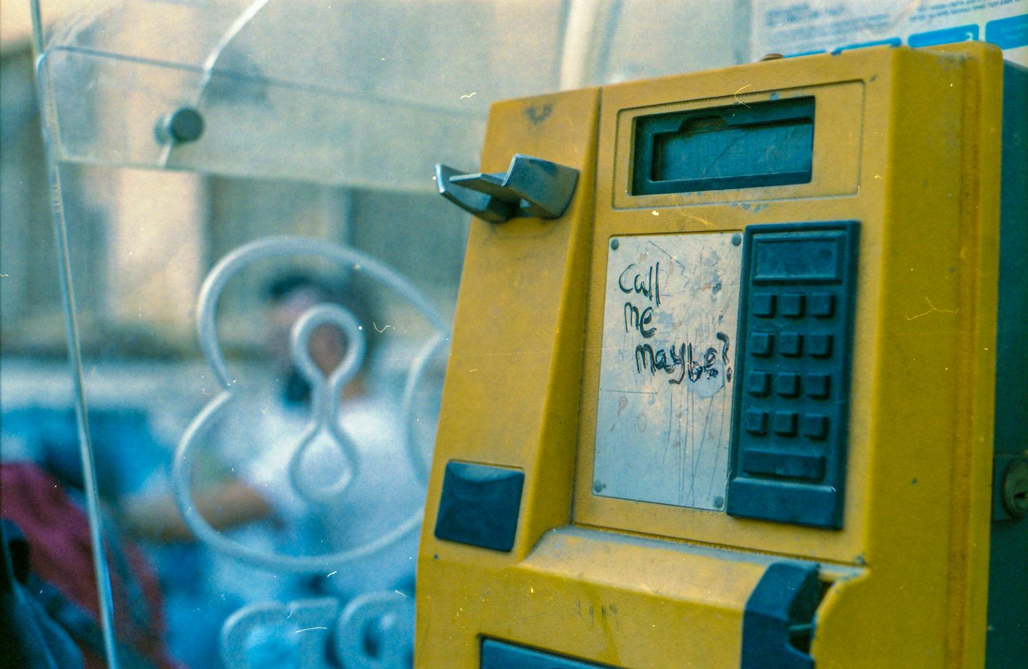 Yellow public payphone with graffiti sticker and numeric keypad, mounted inside a glass telephone booth.
