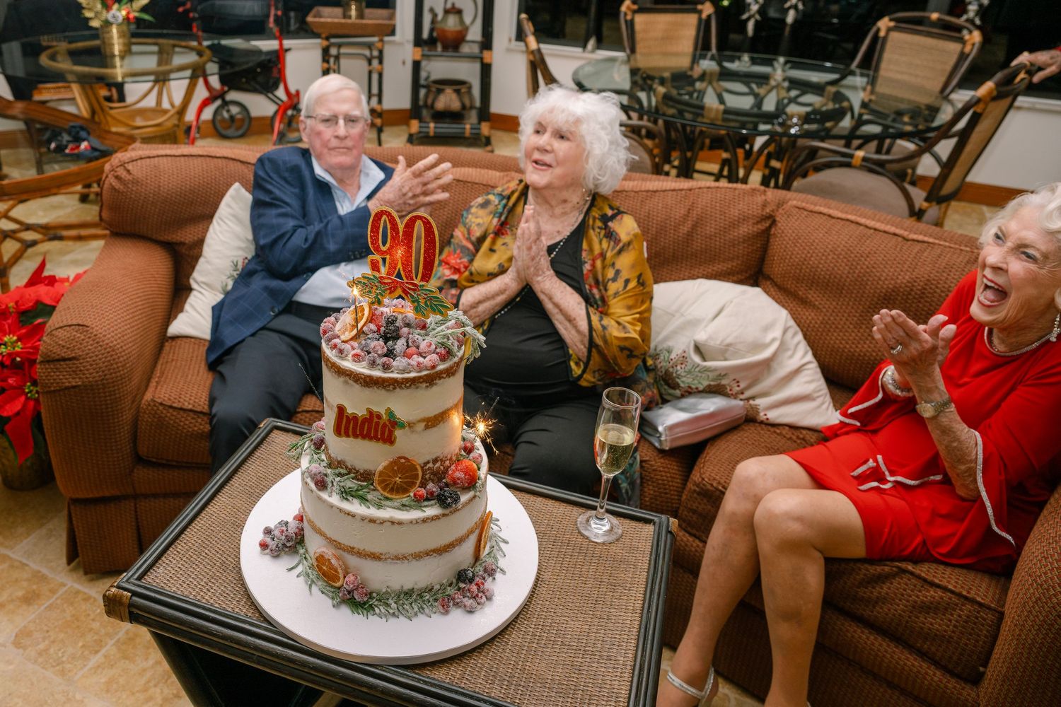 People gathered in a living room celebrating with a birthday cake and decorations for a 90th birthday party.