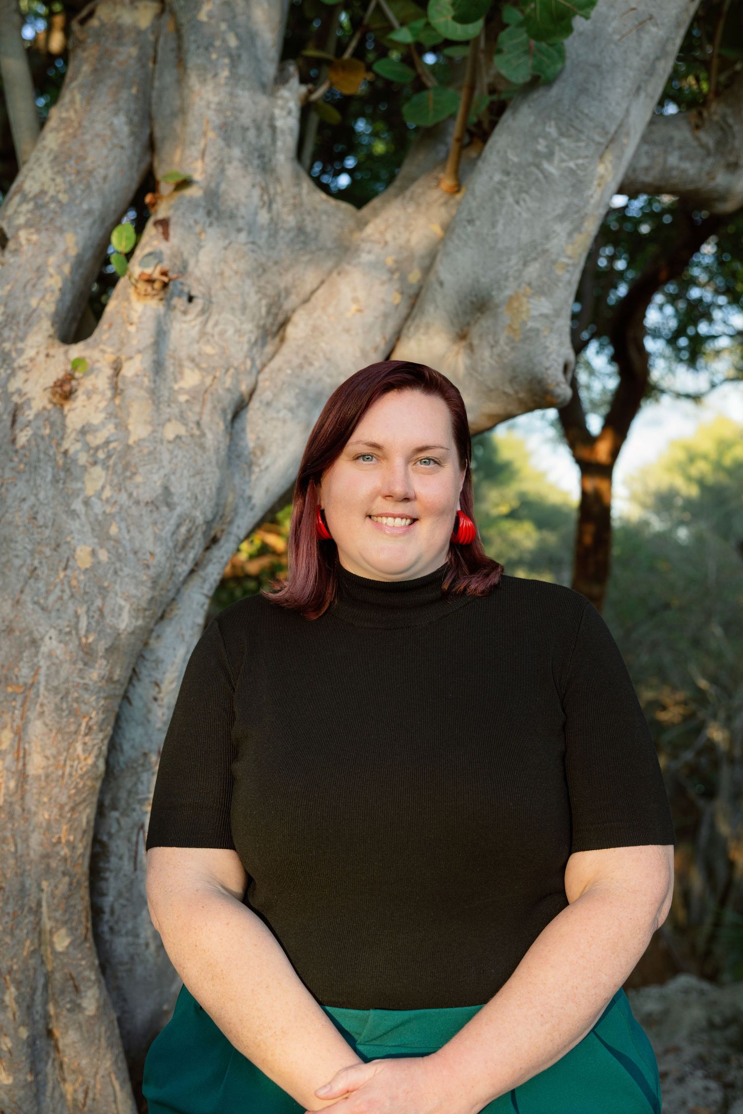 A person in a black top and green skirt poses against a backdrop of tree trunks in natural outdoor lighting.