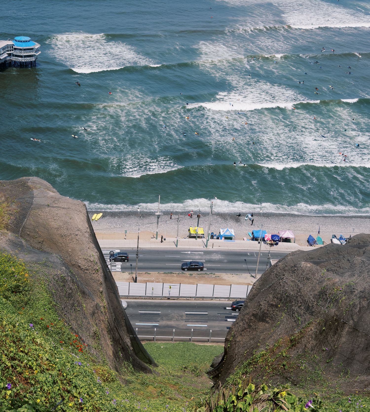 Aerial view of a coastal highway along a sandy beach with colorful umbrellas, ocean waves, and a pier in Lima, Peru.