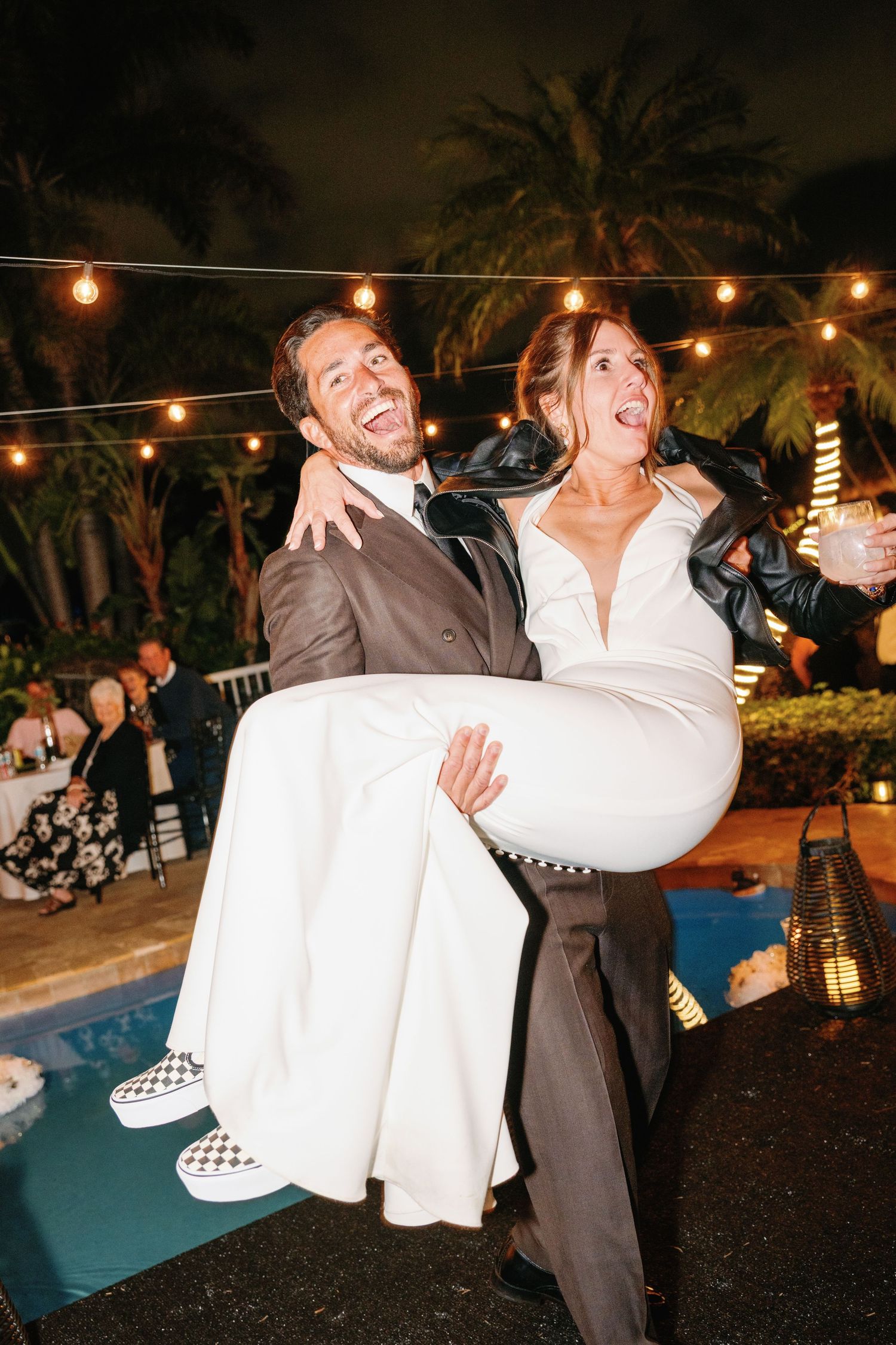 A romantic wedding couple shares playful moments during their reception under string lights at a tropical outdoor venue.
