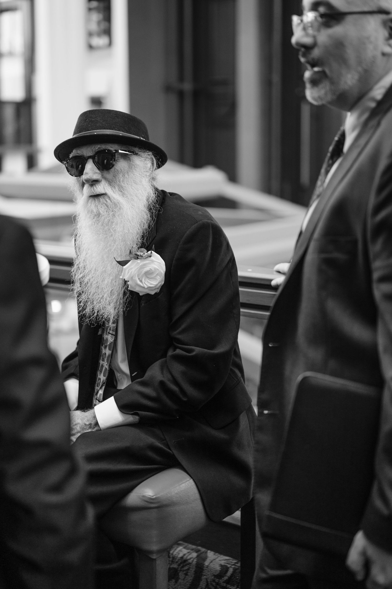 Elderly man with long white beard, sunglasses, and fedora hat sits outdoors in a black suit with a white rose.