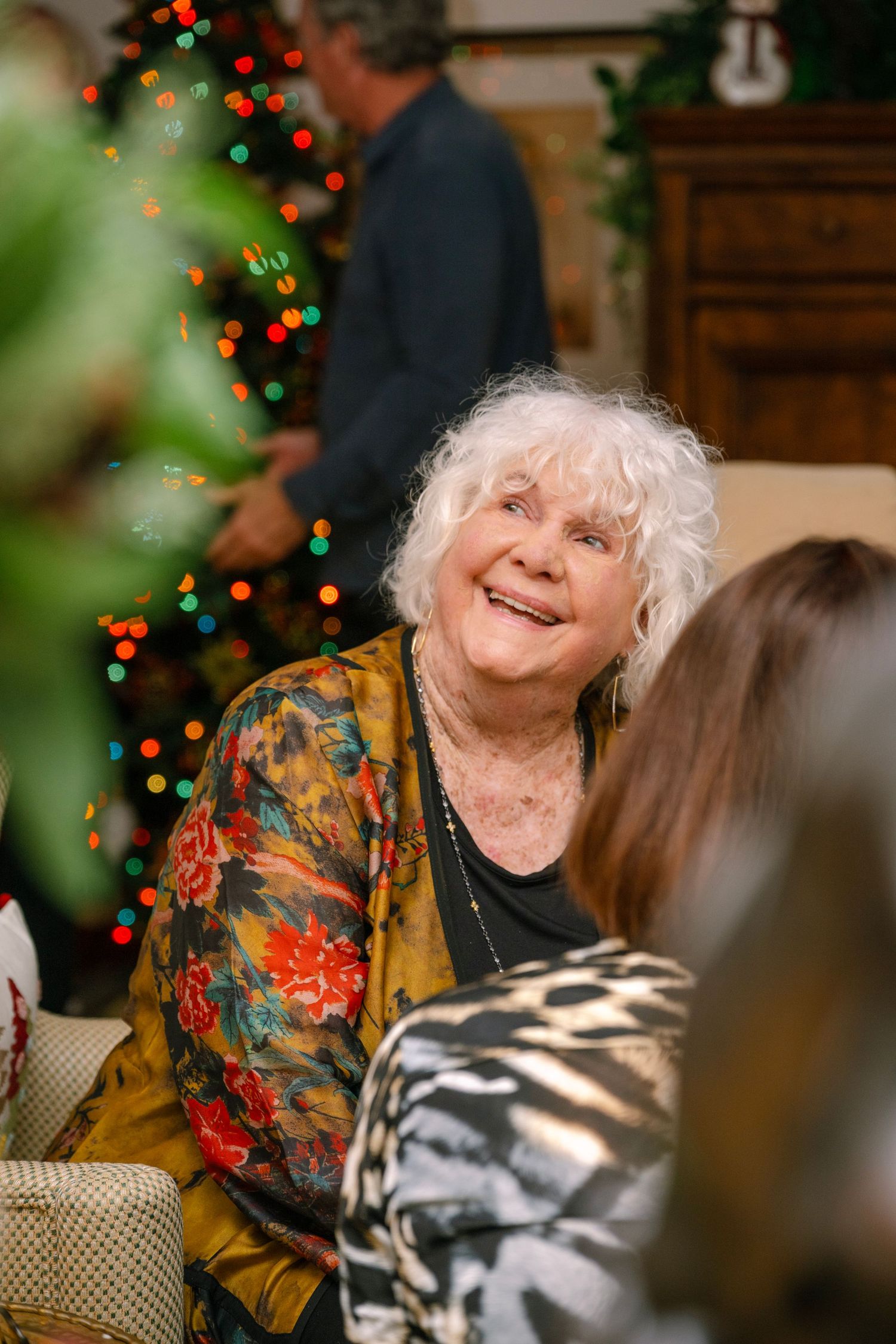 Elderly person in colorful floral top smiling warmly during Christmas celebration with festive lights in background.