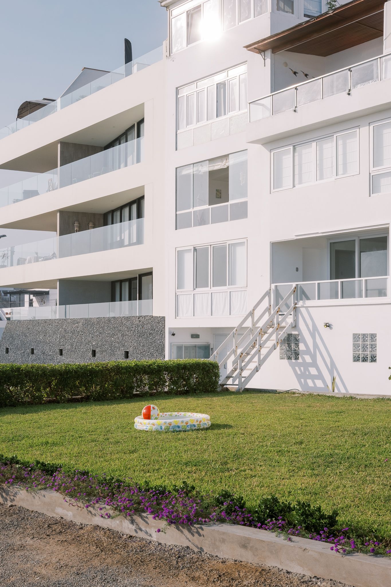 White modern apartment building with balconies, green lawn, small inflatable pool, and purple flowers on a sunny day.