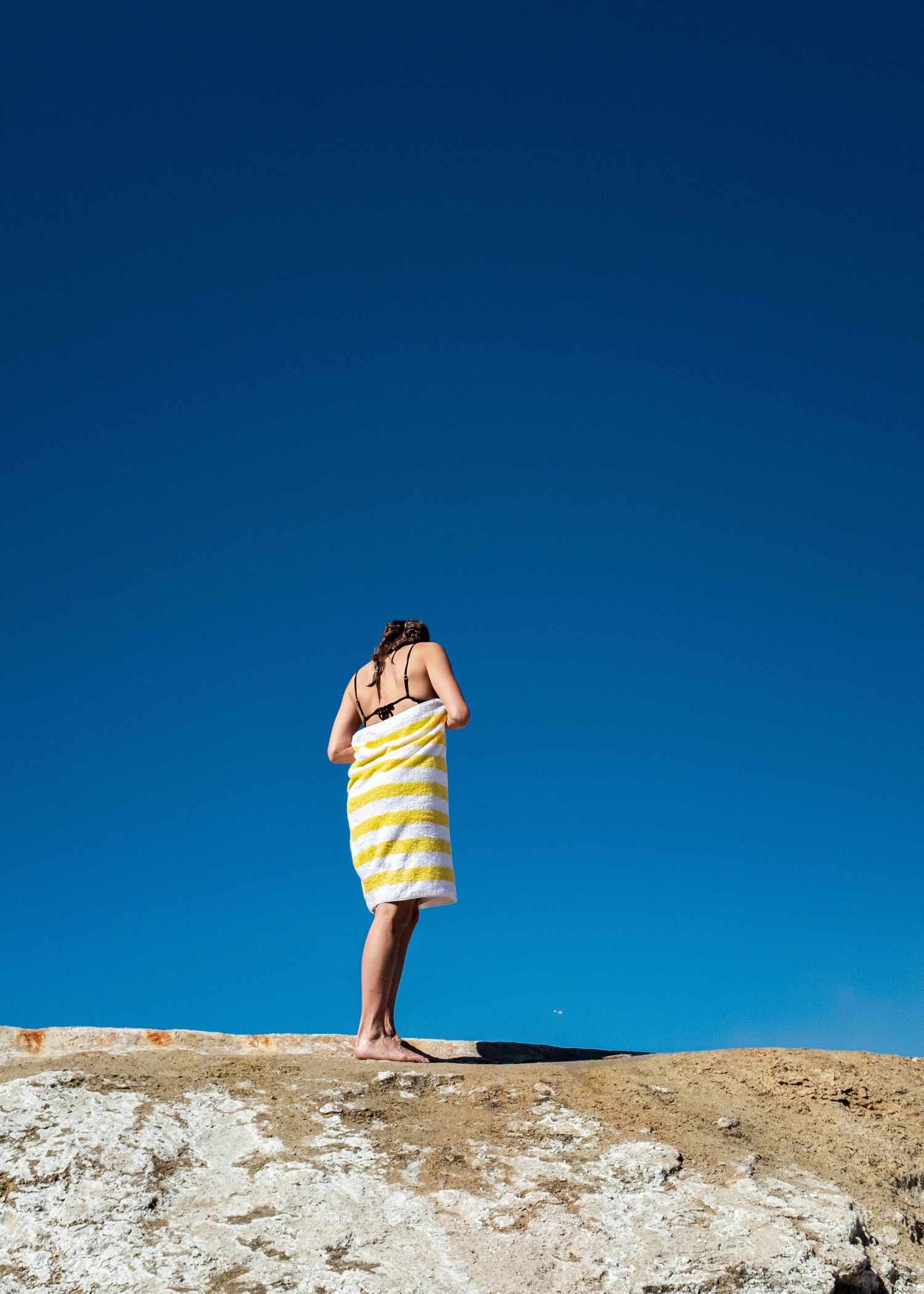 Woman wrapped in yellow striped towel standing on rocky terrain against a clear blue sky.