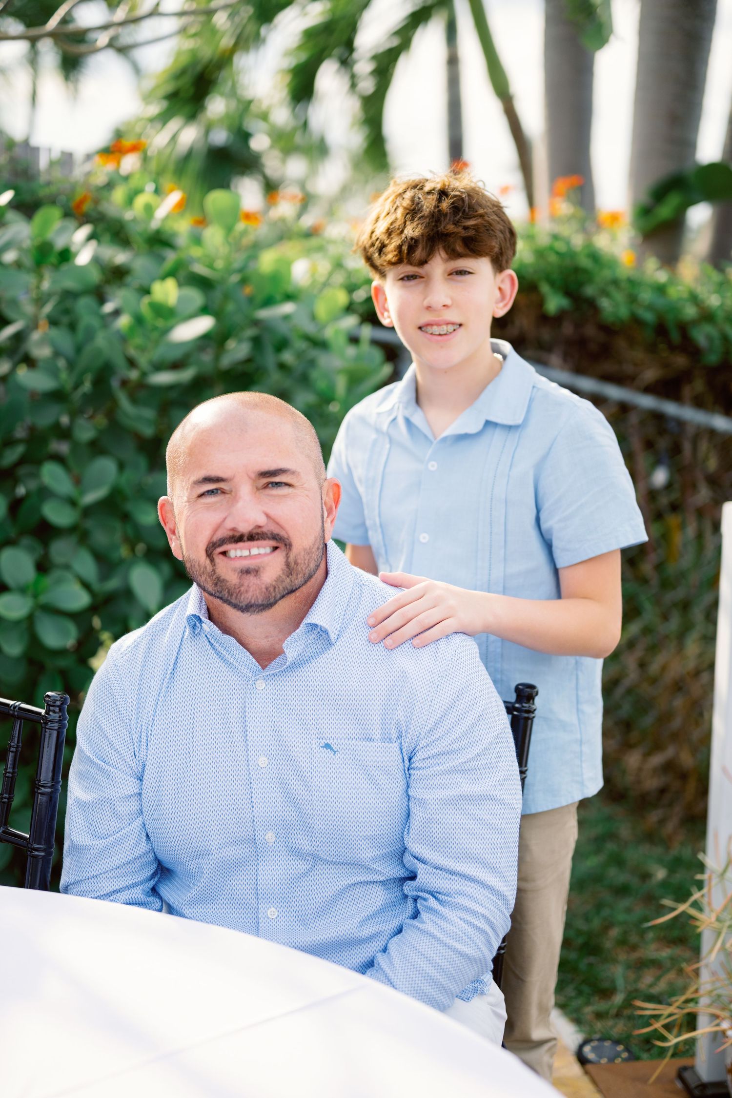 Two people in light blue shirts pose together in a lush garden setting.