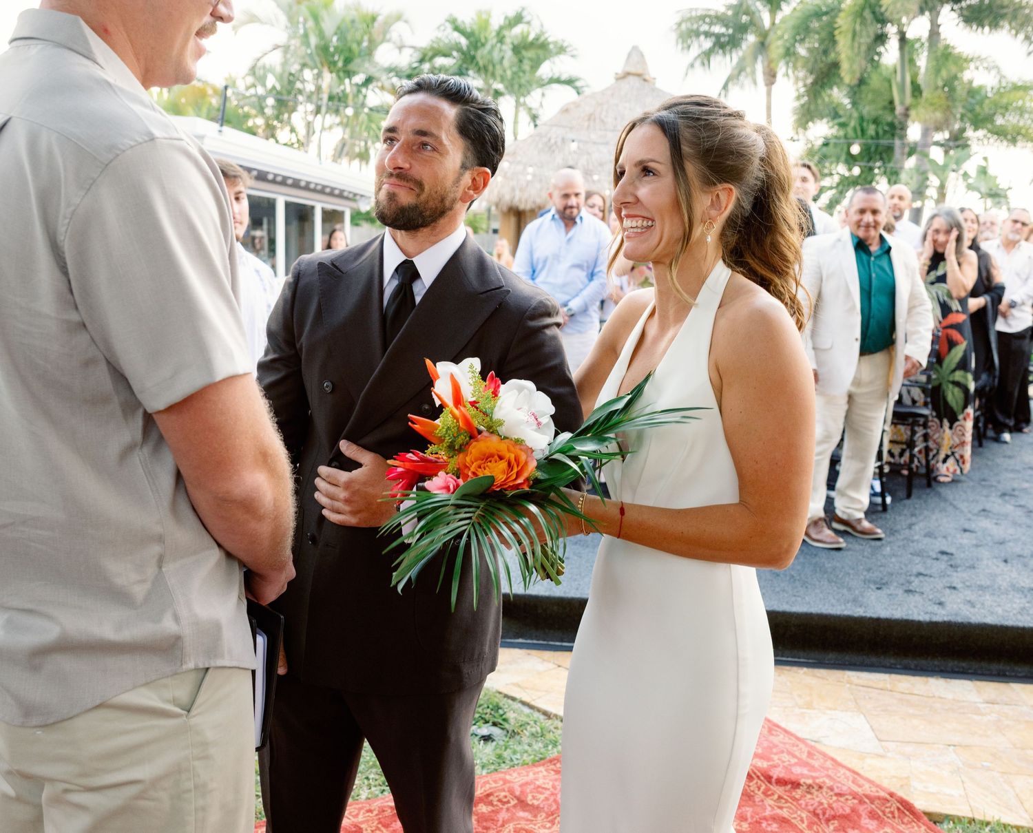 A couple shares a joyful moment during an outdoor wedding ceremony with palm trees in the background.