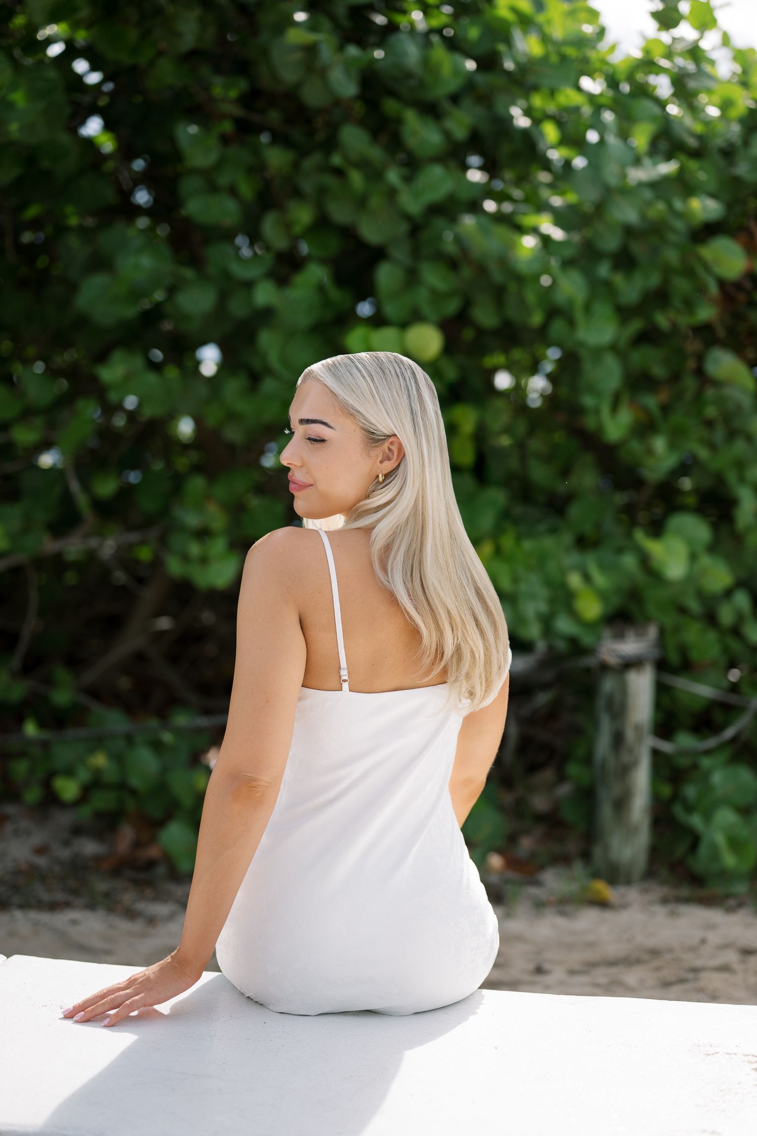 A person in a white tank top sits on a beach with lush green foliage in the background.