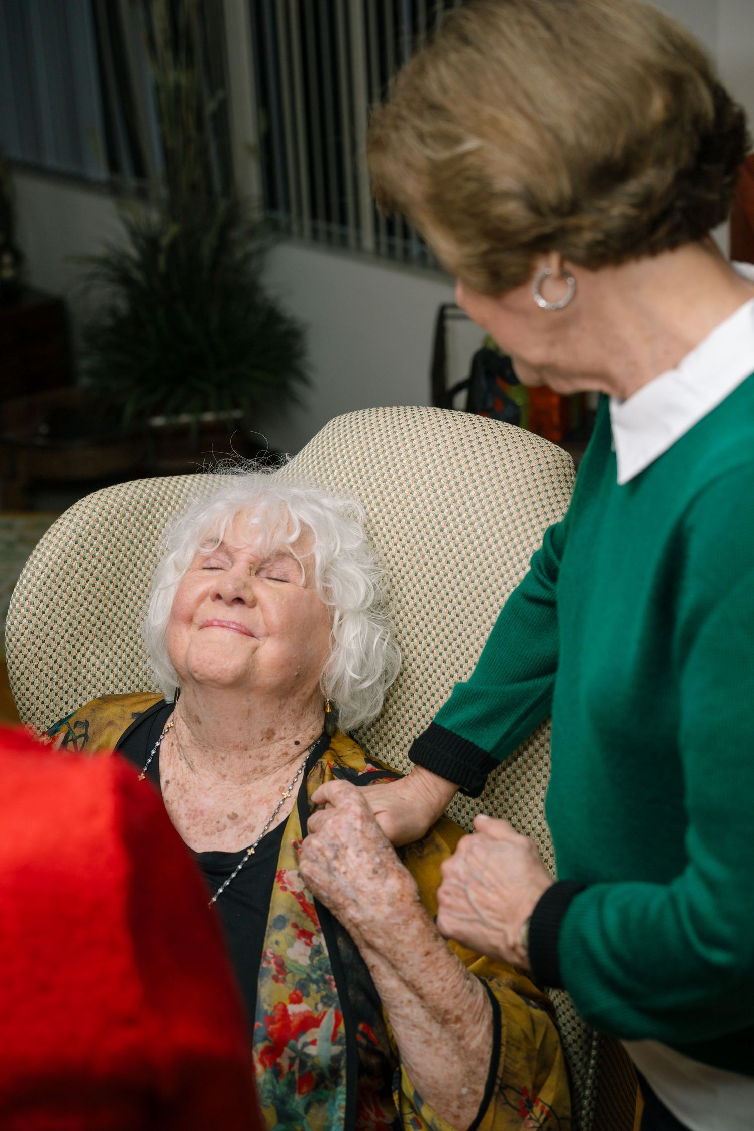 Two people share a tender moment in a living room with a cream colored chair and red accent pillow.