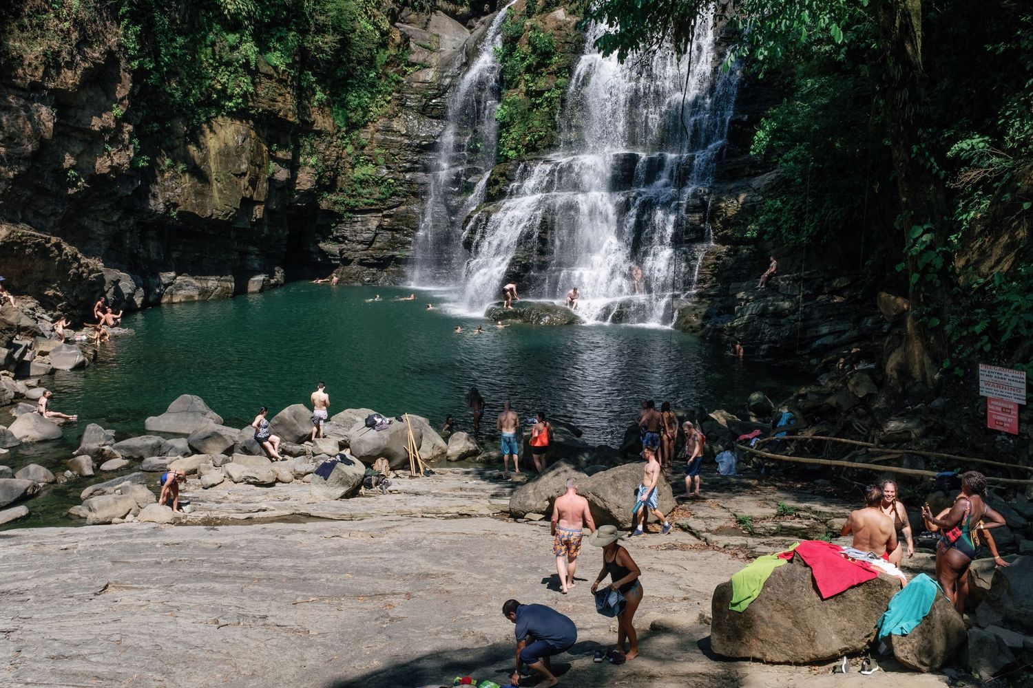 Tourists relaxing on rocks beside a lush tropical waterfall flowing into a green pool surrounded by dense jungle.