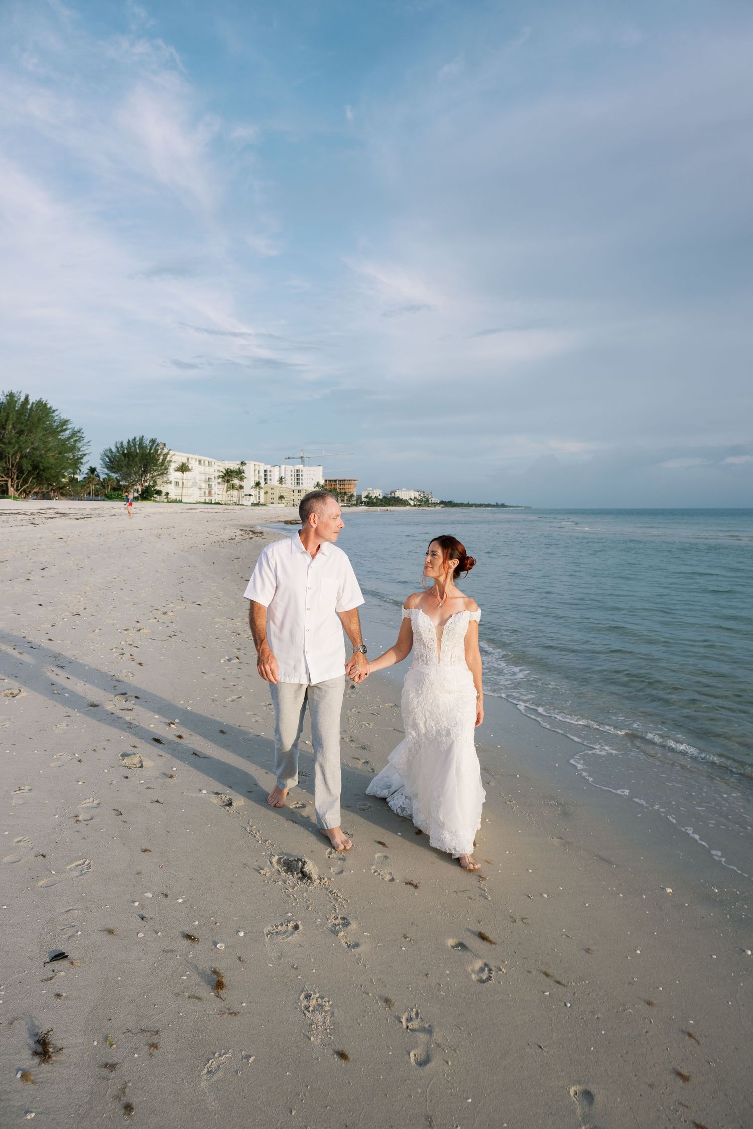 A couple in white attire walks hand in hand along a pristine beach shoreline with blue water and sky in the background.
