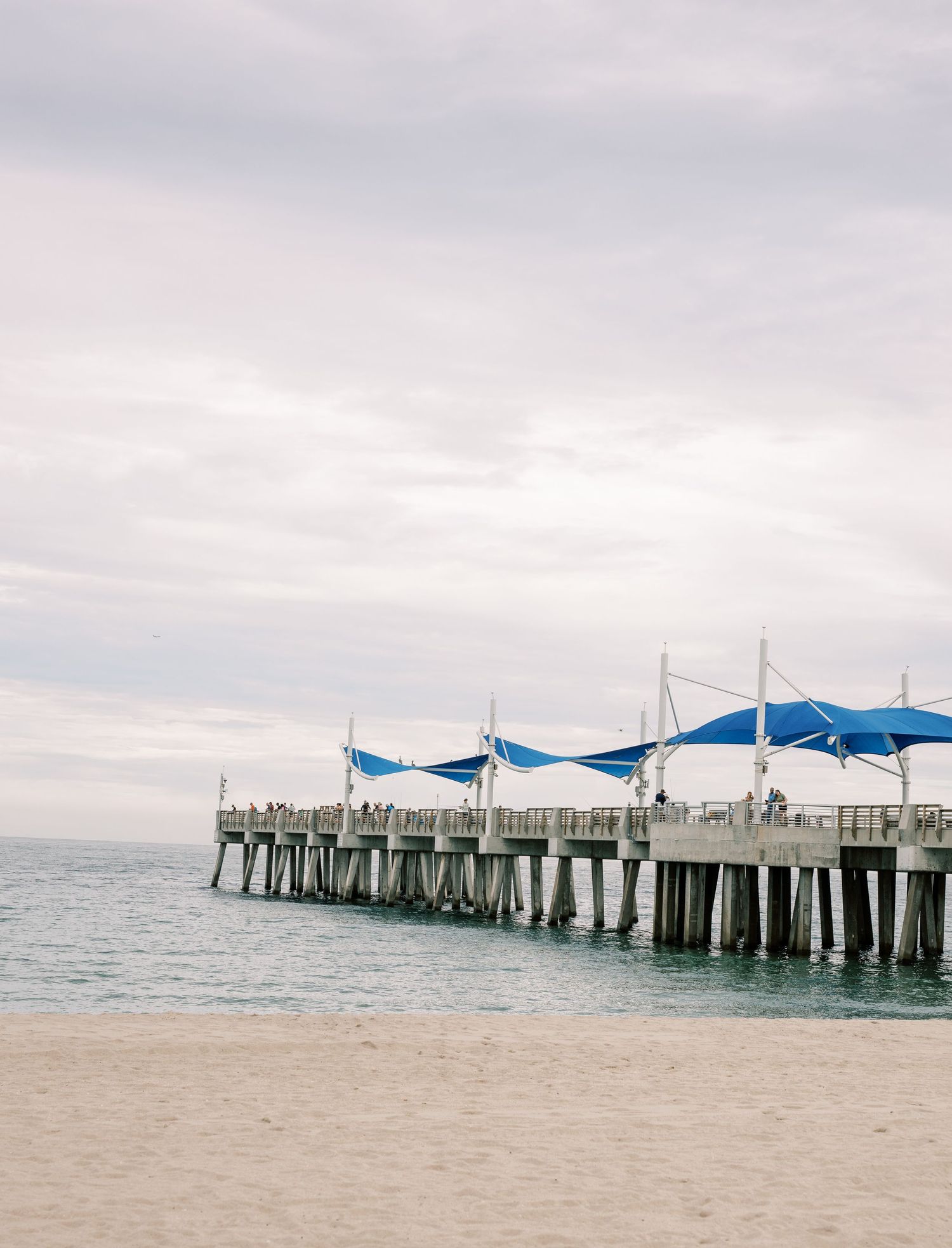 Wooden pier with blue canopies extends into the ocean under an overcast sky.