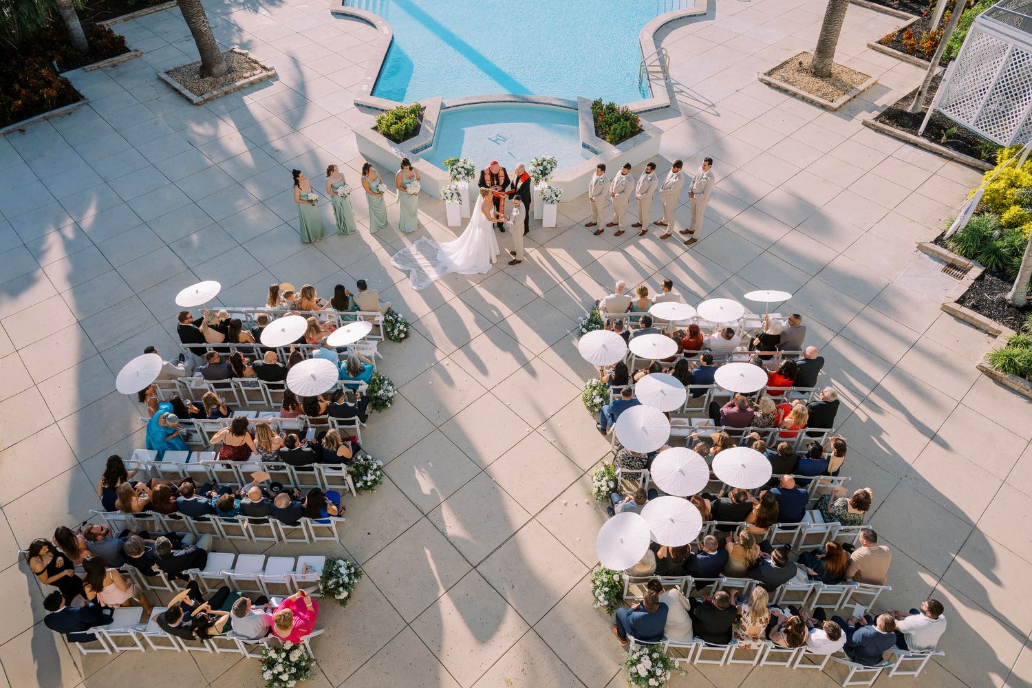 Aerial view of people dining at outdoor tables with white umbrellas on a sunny patio or plaza.