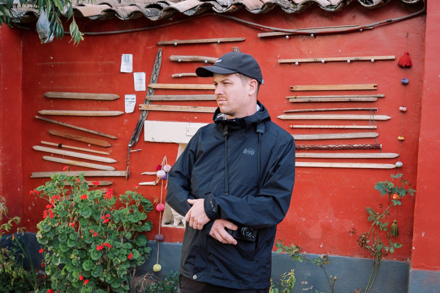 Two people in dark jackets stand near a red wall with tools and gardening equipment mounted on it.