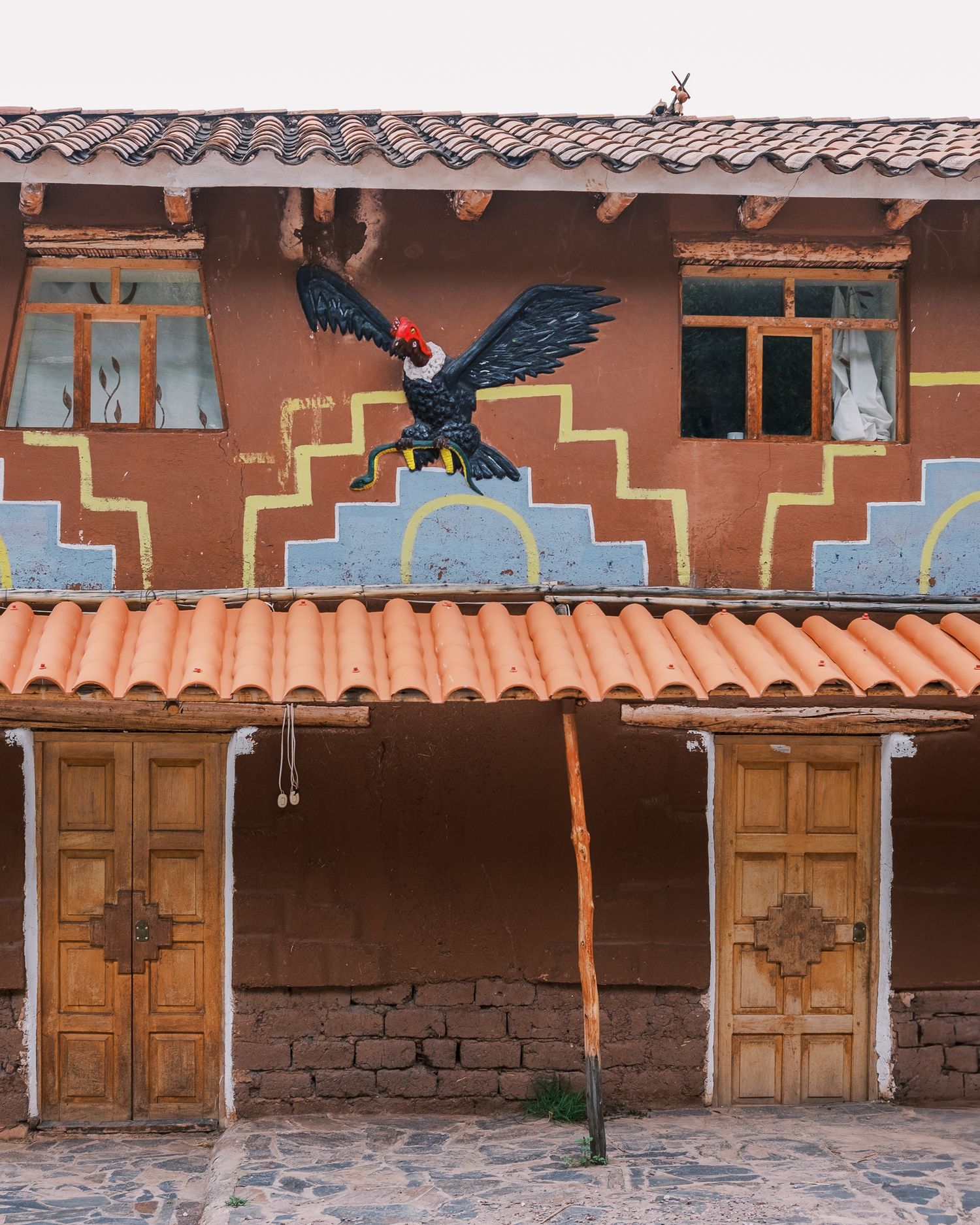 Colorful Andean building with terracotta roof tiles, wooden doors, and a painted condor mural on the adobe wall.