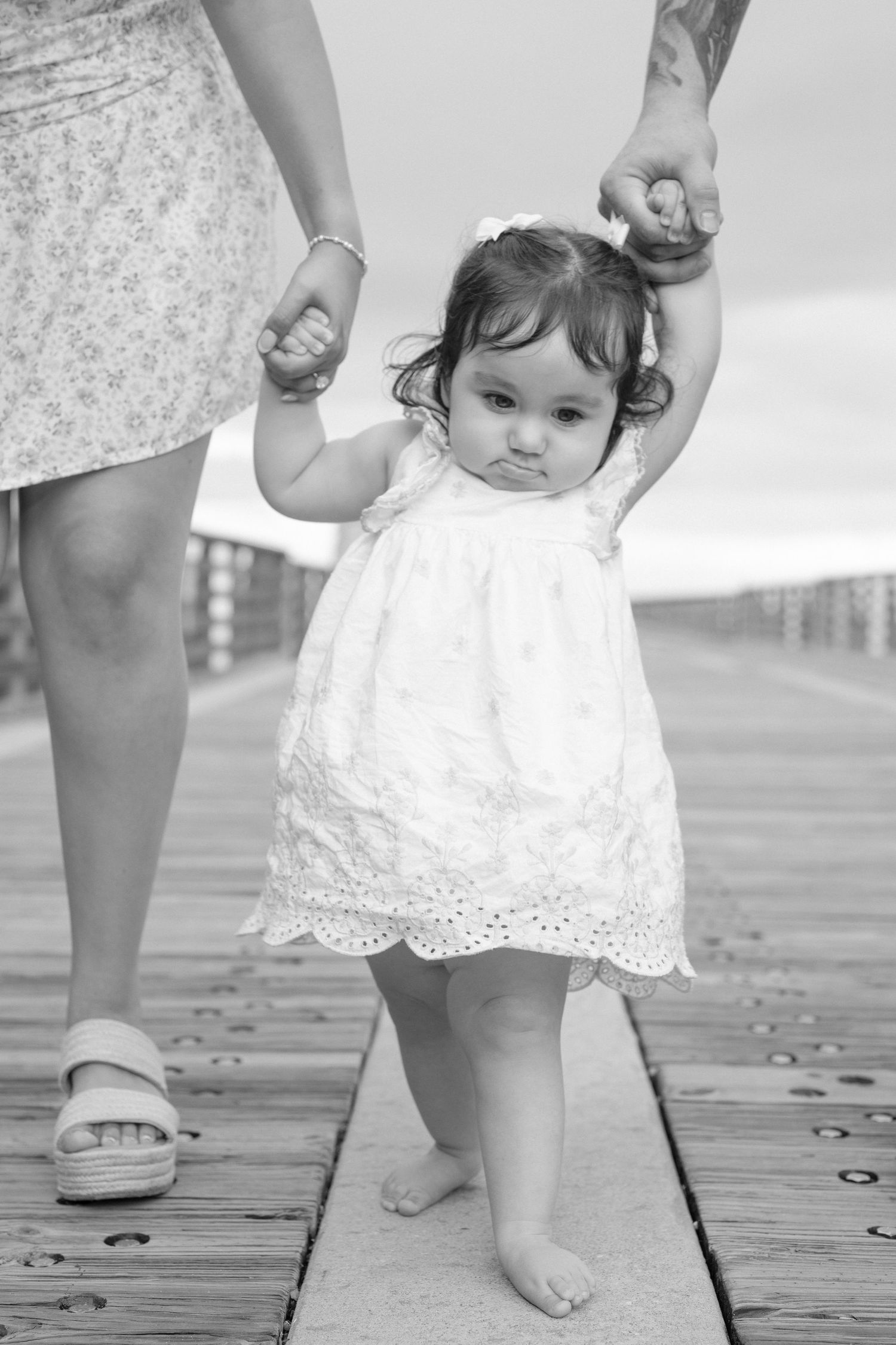 A baby in a white lace dress takes first steps on a wooden boardwalk while holding hands with an adult.