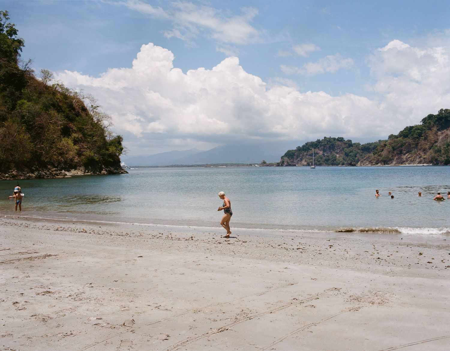 Tropical beach scene with calm blue water, lush green cliffs, white clouds, and people enjoying the sandy shore.