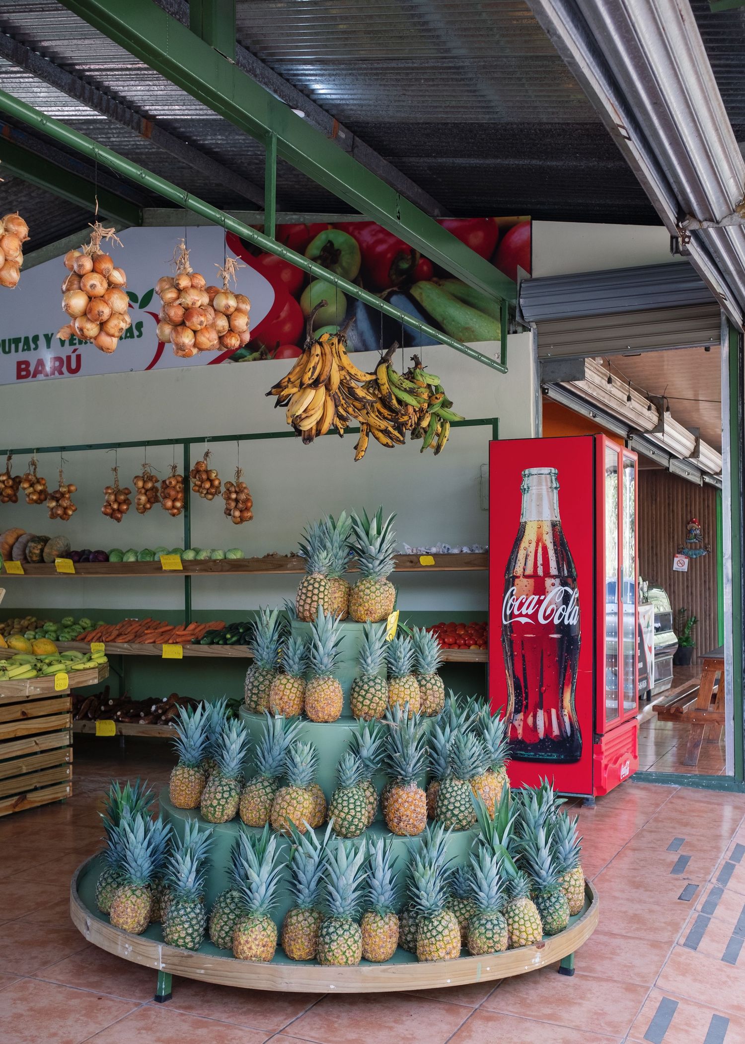 Tropical fruit market displaying a pyramid of fresh pineapples, hanging bananas, onions, and a Coca-Cola refrigerator in background.