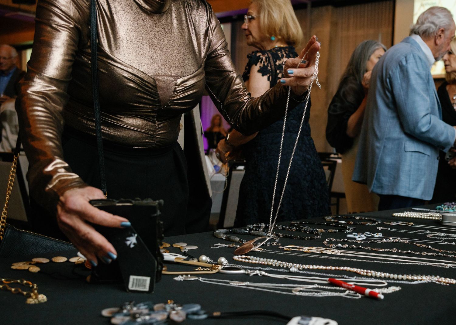 Hands arranging jewelry pieces on a dark display table at a crowded event or marketplace.