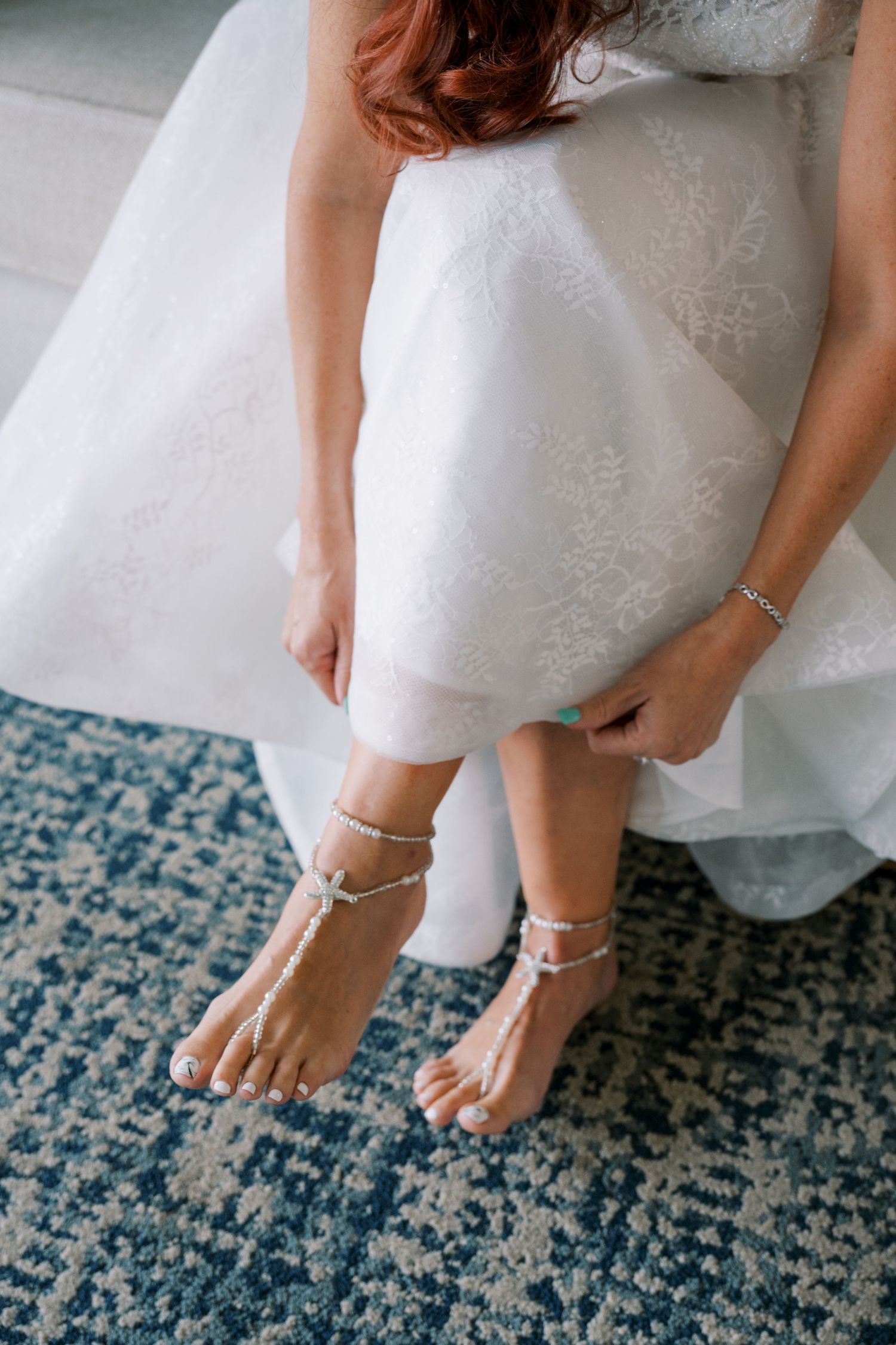 Close-up view of bride's feet wearing delicate ankle straps and white wedding shoes on a blue patterned carpet.