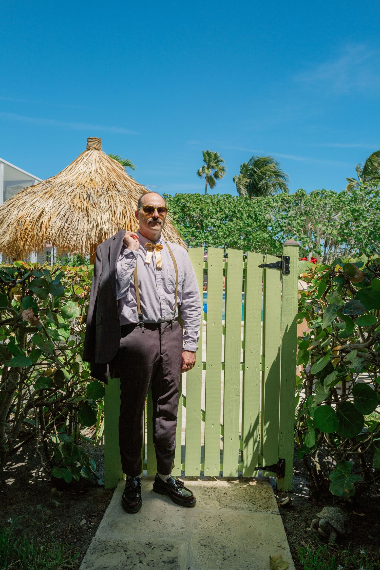 Someone stands in front of a green wooden gate with a tiki umbrella and tropical plants in a sunny backyard setting.