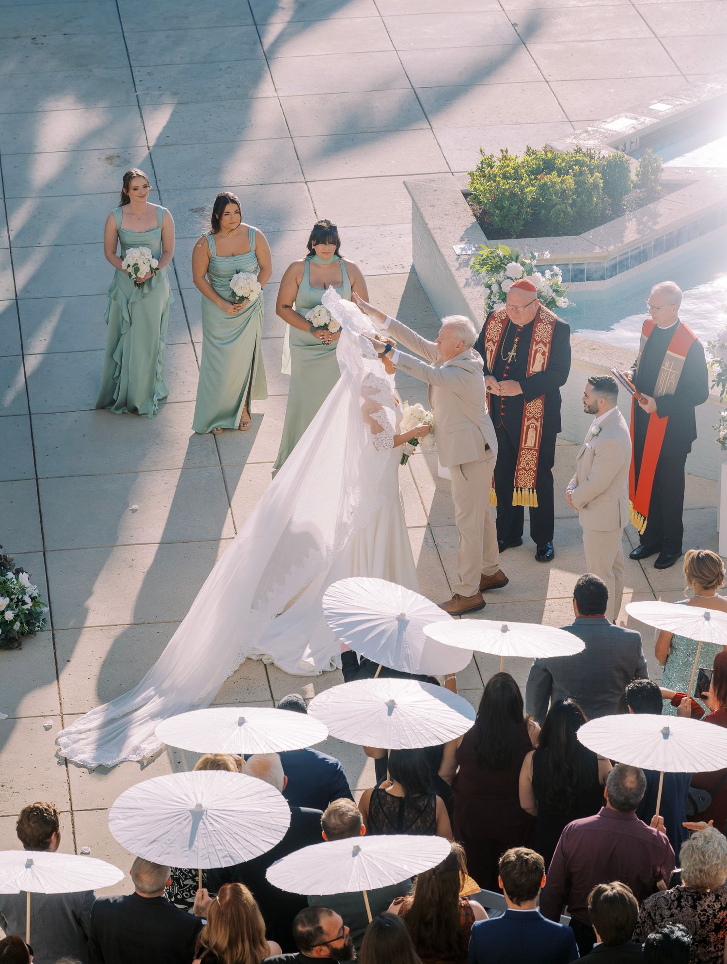 Wedding party stands in line with white parasols creating an elegant outdoor ceremony backdrop.