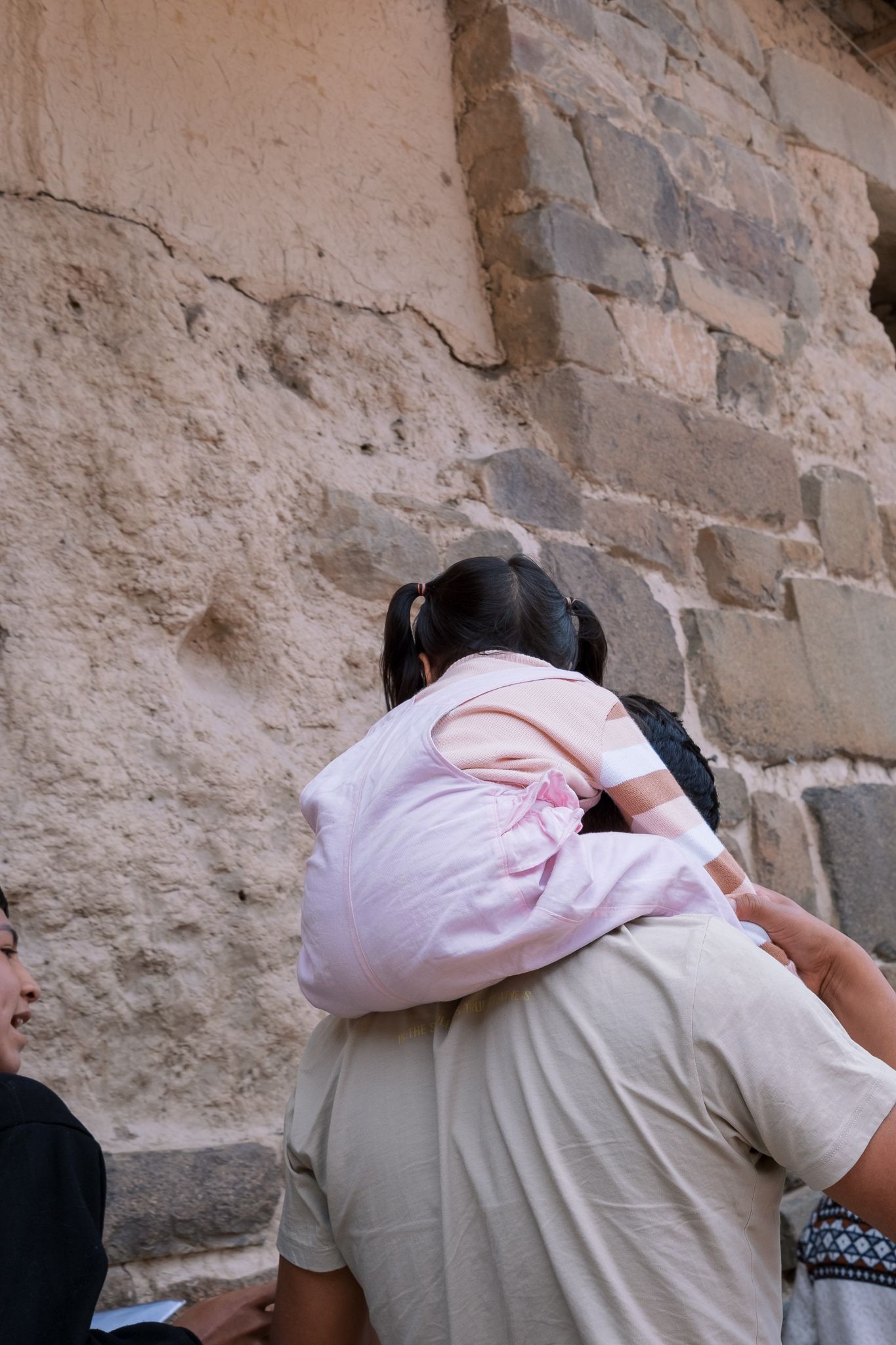 A young girl in a pink outfit sits on an adult's shoulders facing an ancient stone wall.