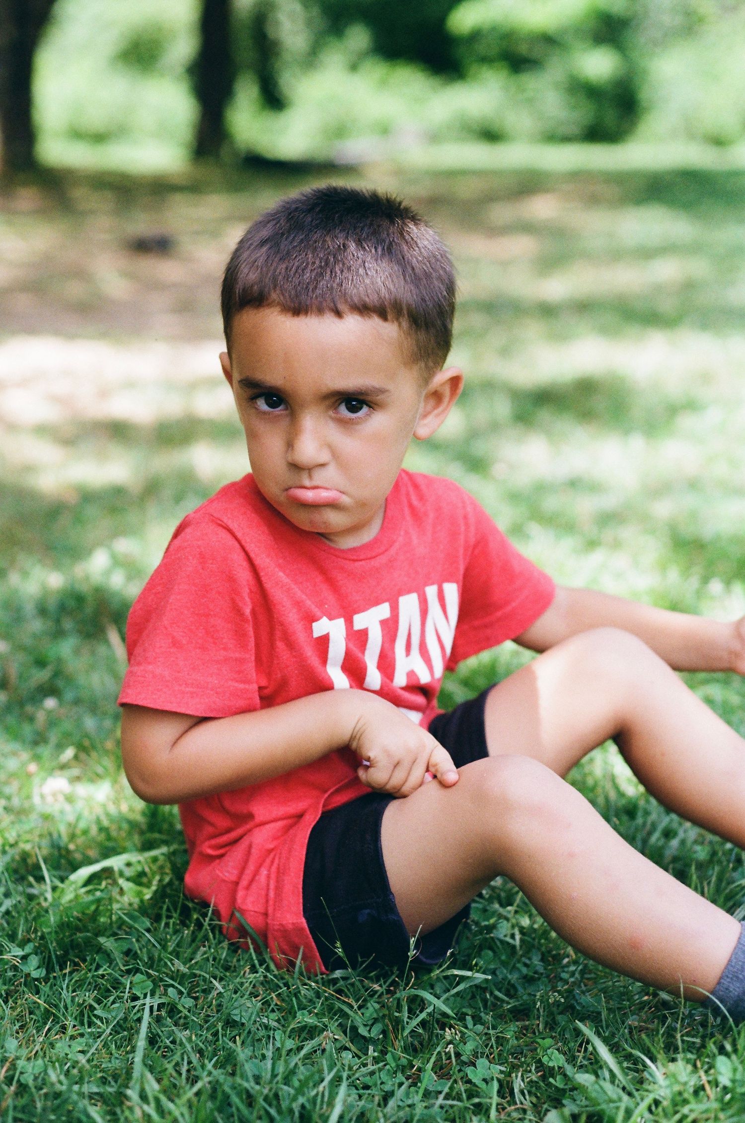 A young boy in a red shirt and black shorts sits on grass in a park, looking at the camera with a pouty expression.