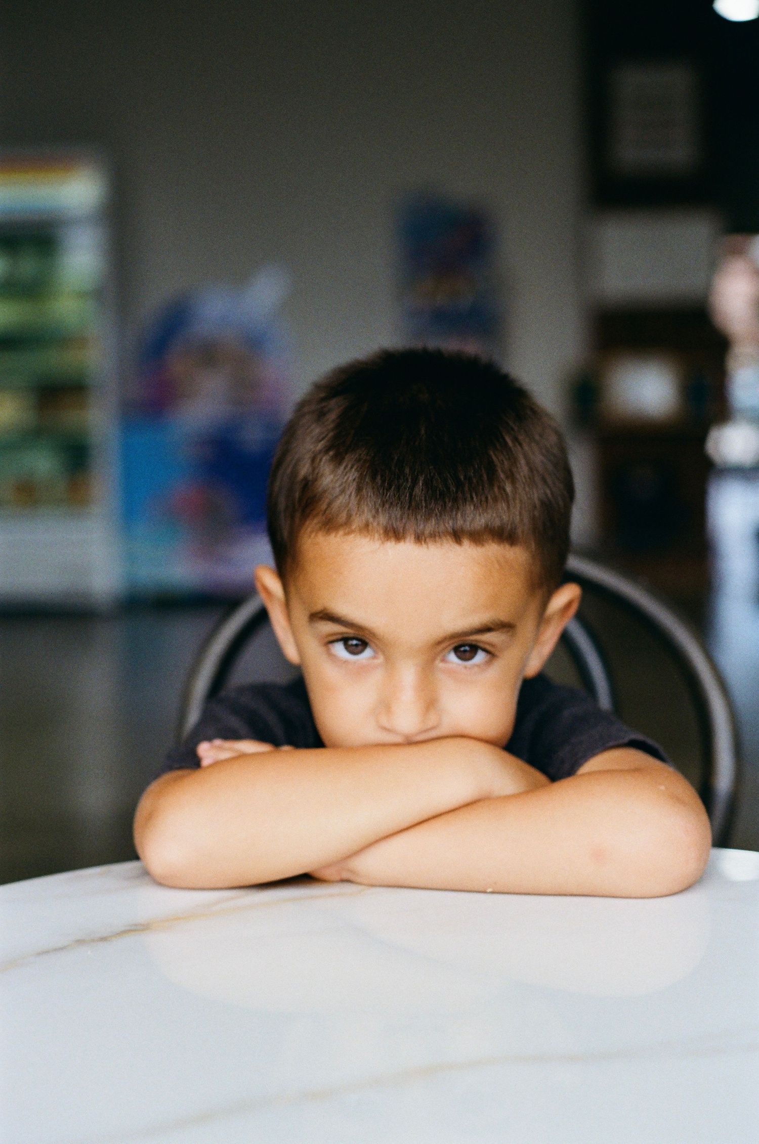 Young boy with crossed arms resting chin on table, looking serious in a casual indoor setting.