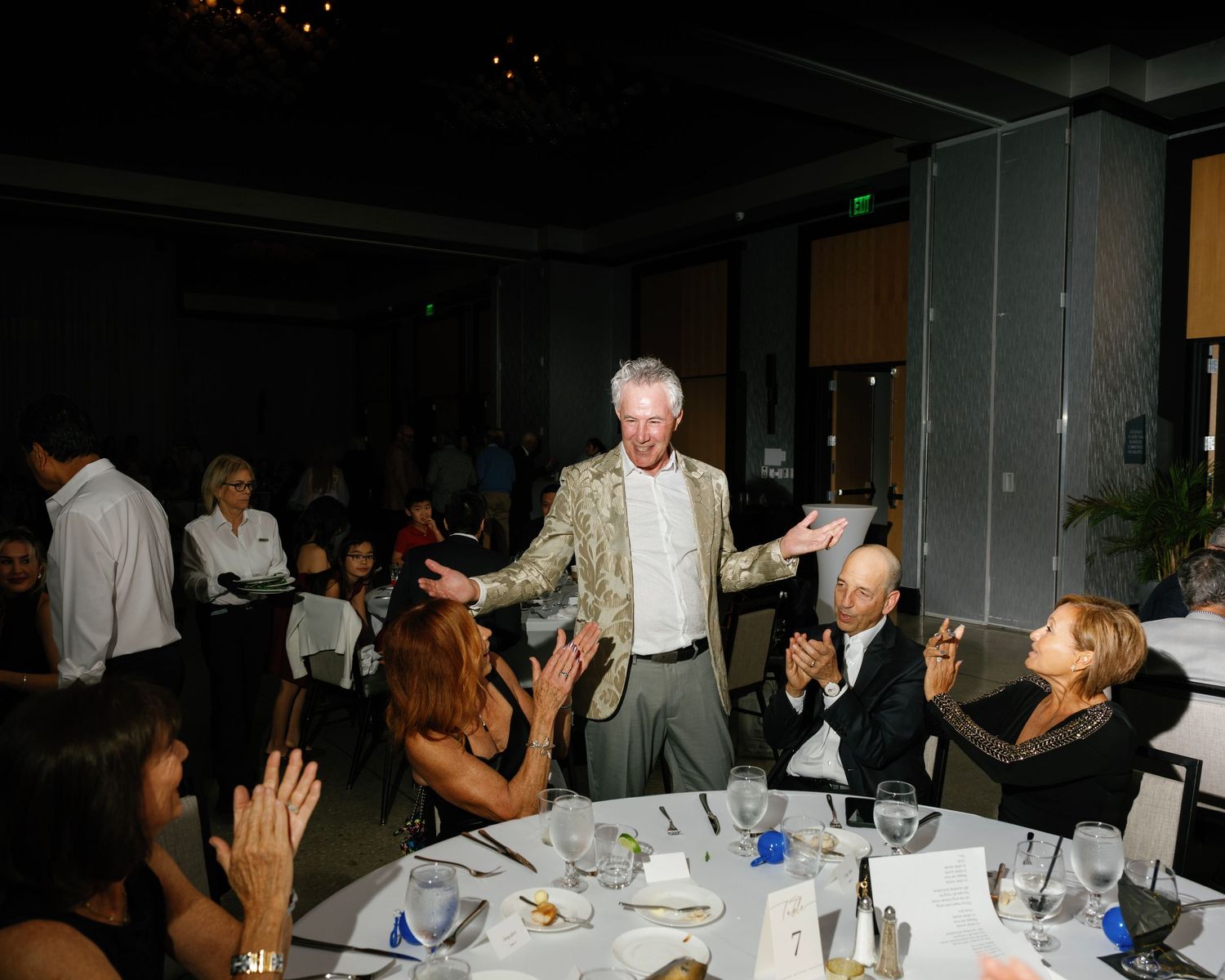 People gathered around tables at an indoor event raising their hands in celebration.