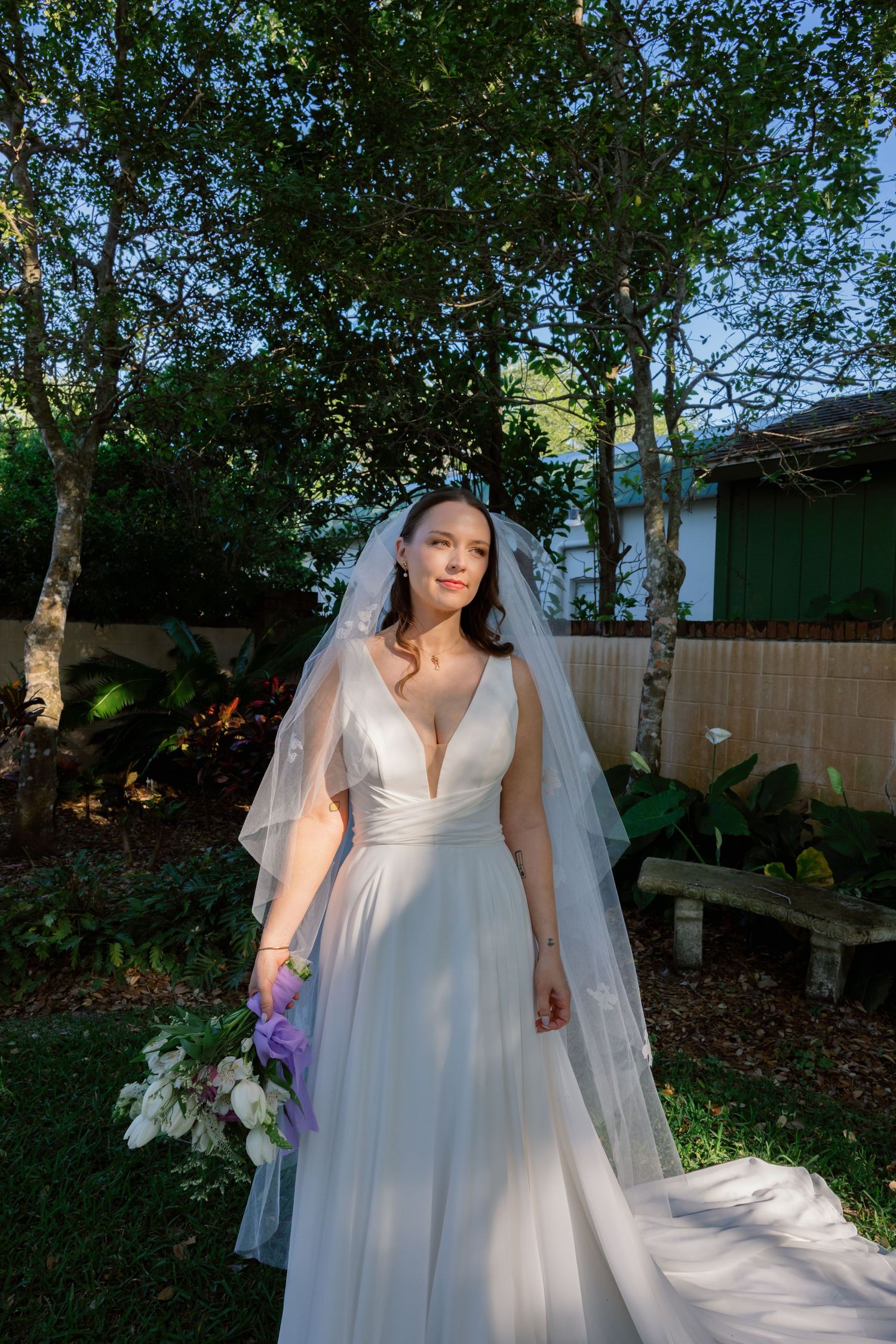 A wedding dress with a flowing veil photographed in a lush garden setting with dappled sunlight.